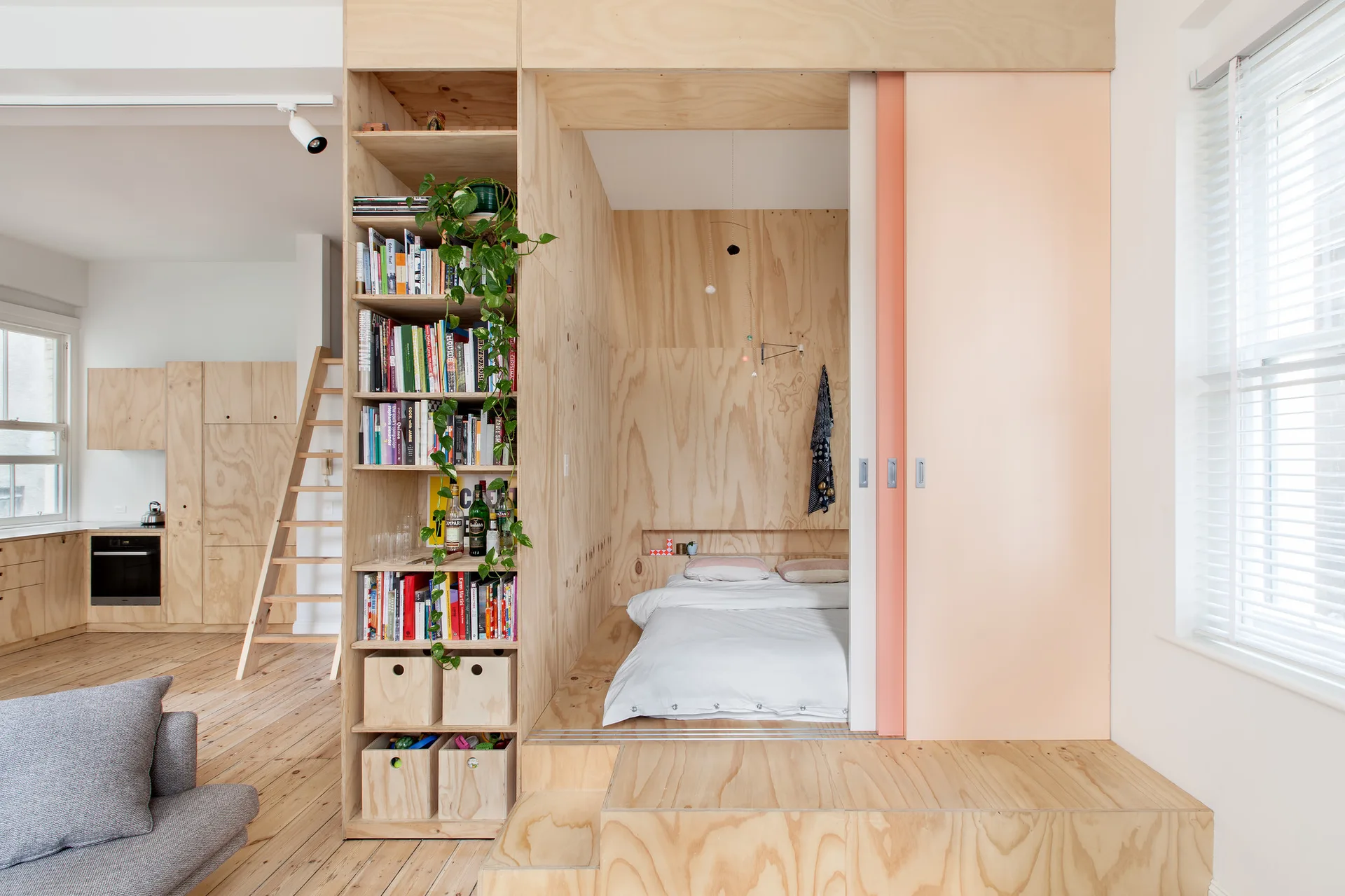 A plywood-framed bedroom structure in a studio apartment features built-in bookshelves, a raised bed nook, and a pale peach sliding door.