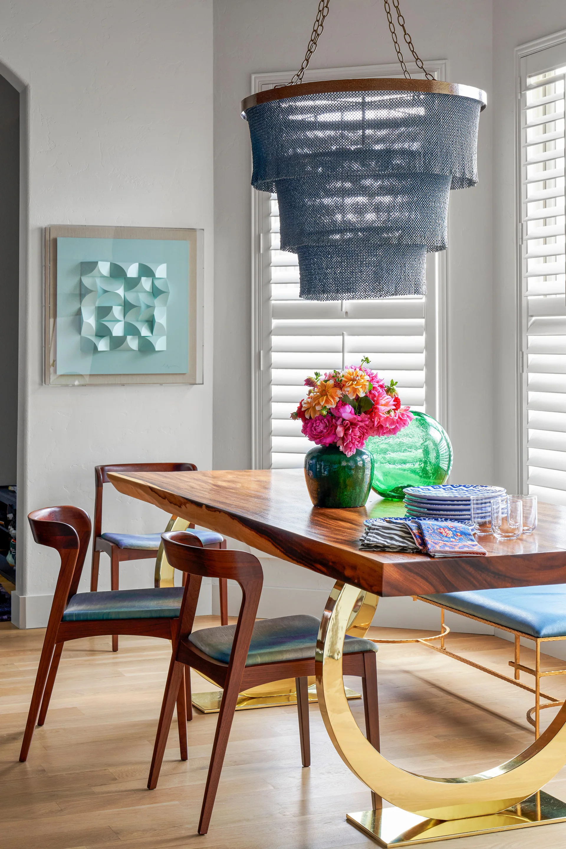 A dining room features a live-edge wood table with a curved brass base, mid-century modern wooden chairs, and a tiered metal mesh pendant light.