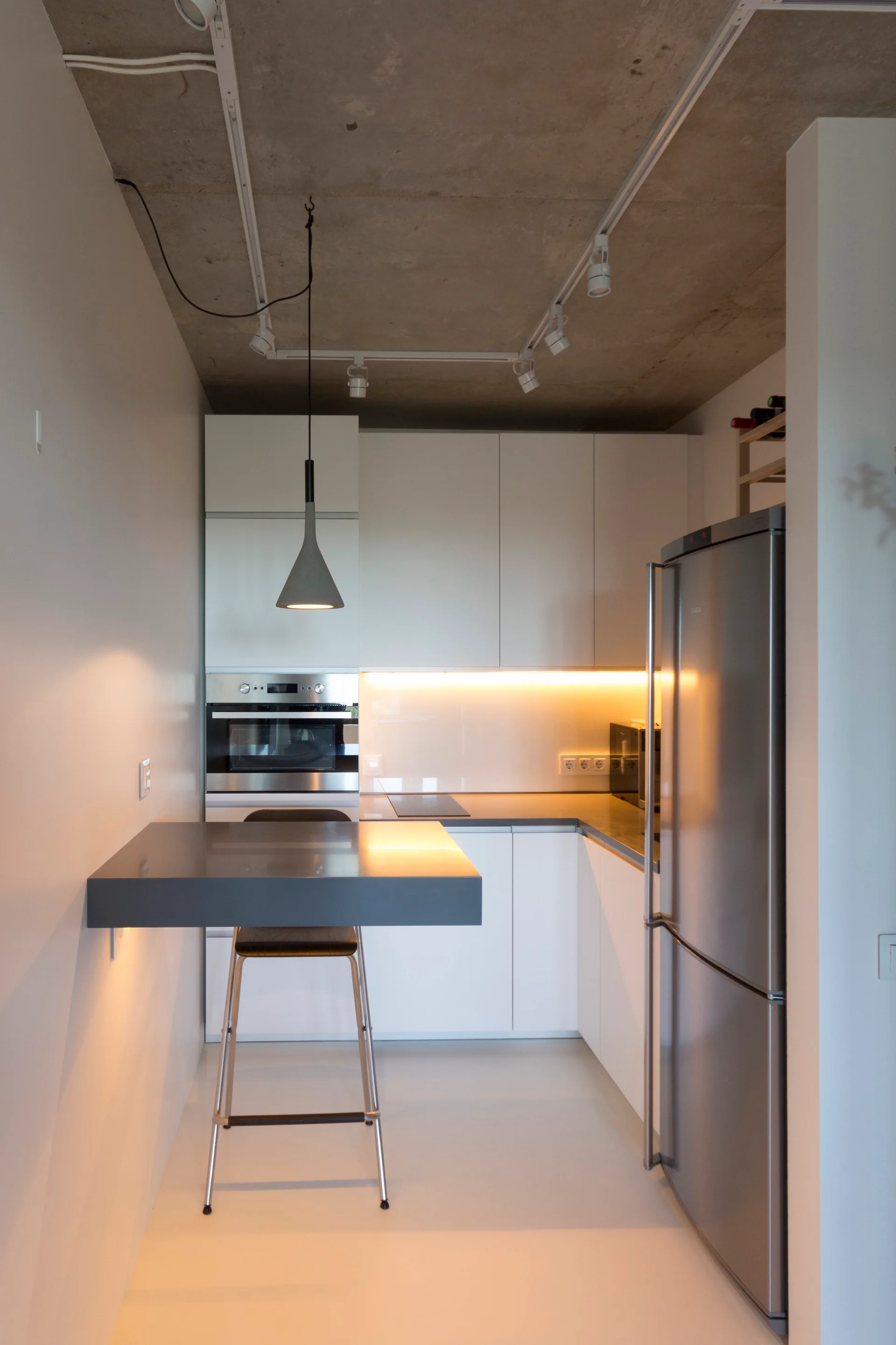 A minimalist kitchen features white cabinets, a stainless steel refrigerator, a grey cantilevered breakfast bar, and an exposed concrete ceiling.