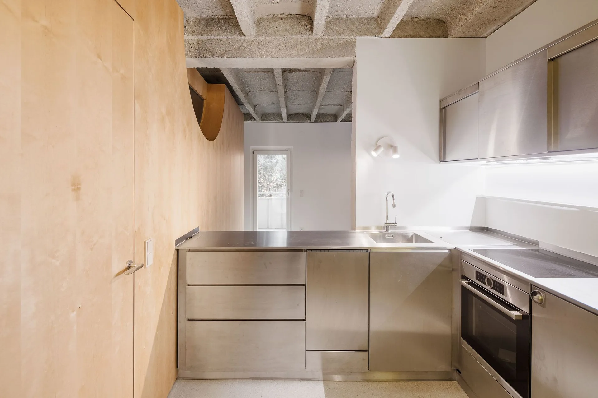 A modern kitchen with stainless steel cabinets and appliances set against a light wood paneled wall under an exposed concrete ceiling.