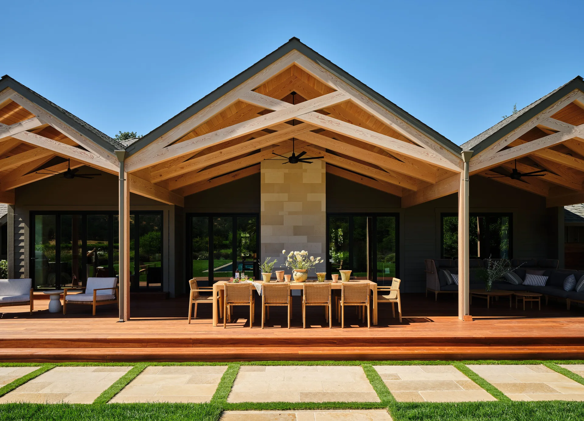 A covered outdoor patio features a wooden dining table under a vaulted timber ceiling alongside a central stone fireplace.
