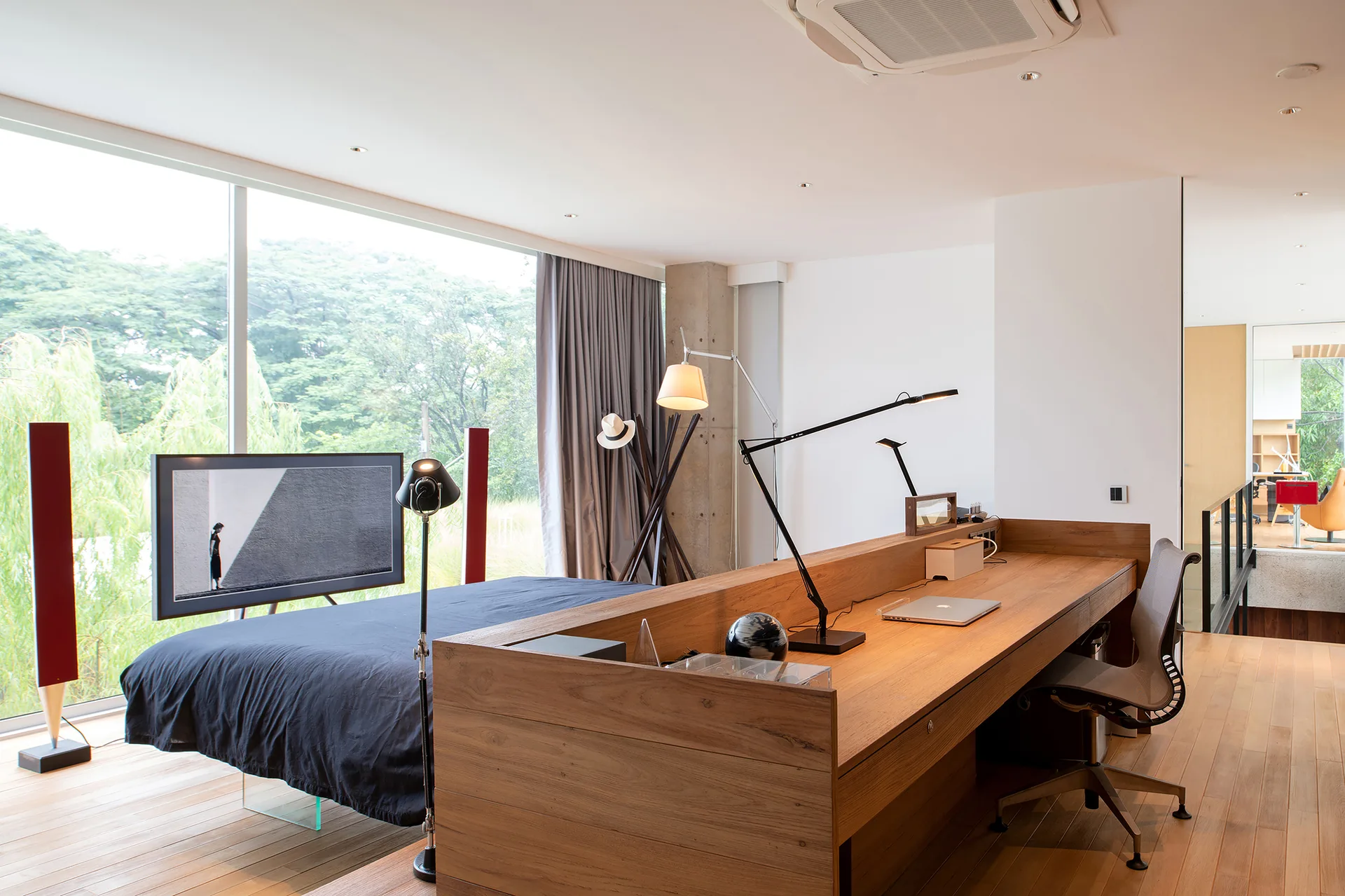 A low wood partition desk stands between a bed and a workspace next to a large floor-to-ceiling window.