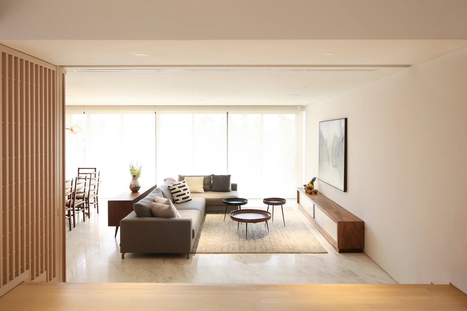 A bright living room features a grey sectional sofa, three small round coffee tables, a low wood media console, and a slatted wood screen.