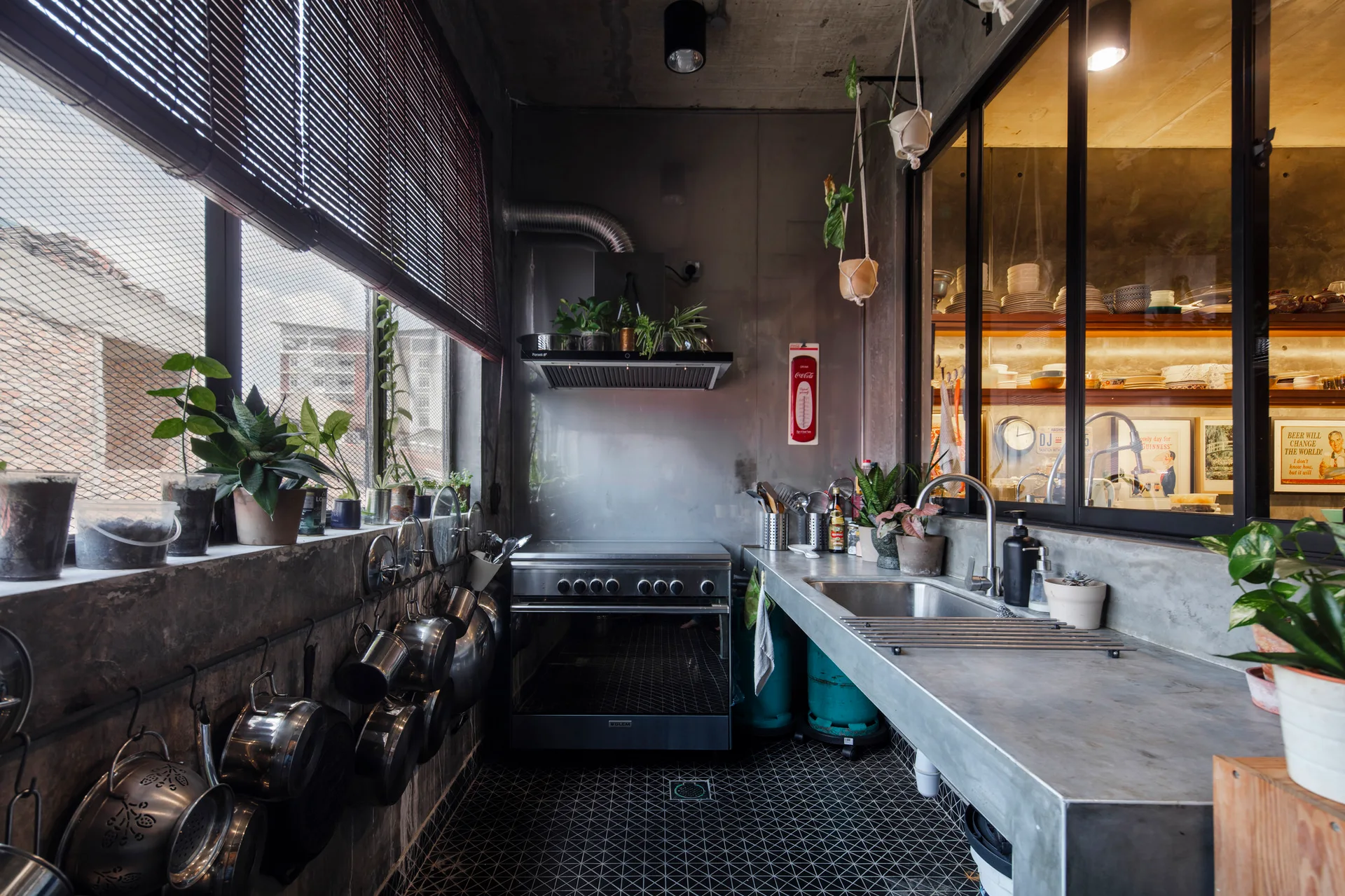 An industrial kitchen featuring raw concrete countertops, a stainless steel stove, hanging pots on a concrete wall, and geometric black-and-white floor tiles.