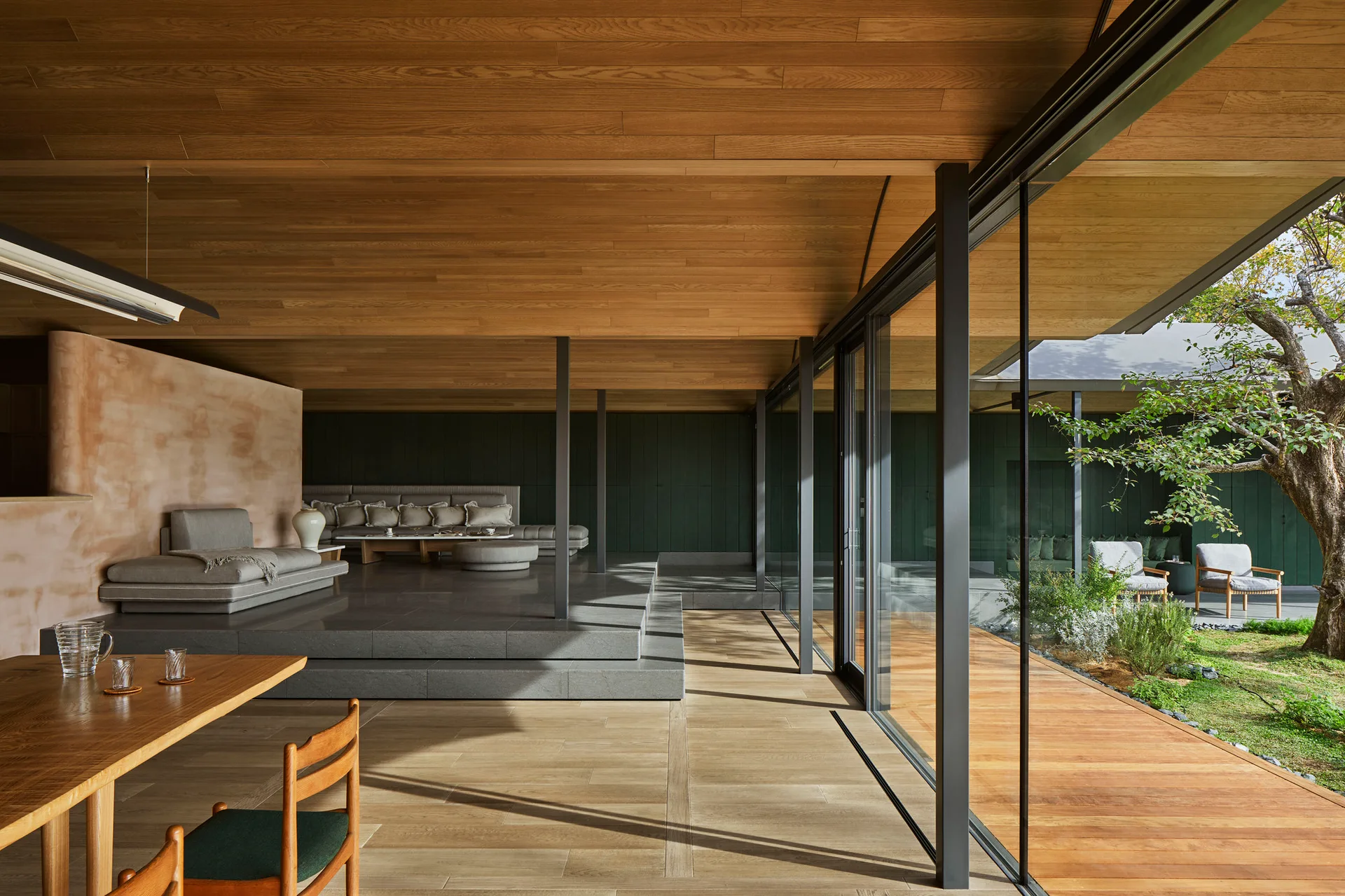 A modern living room features a raised grey stone platform, a natural wood plank ceiling, and large sliding glass walls that open to a garden.