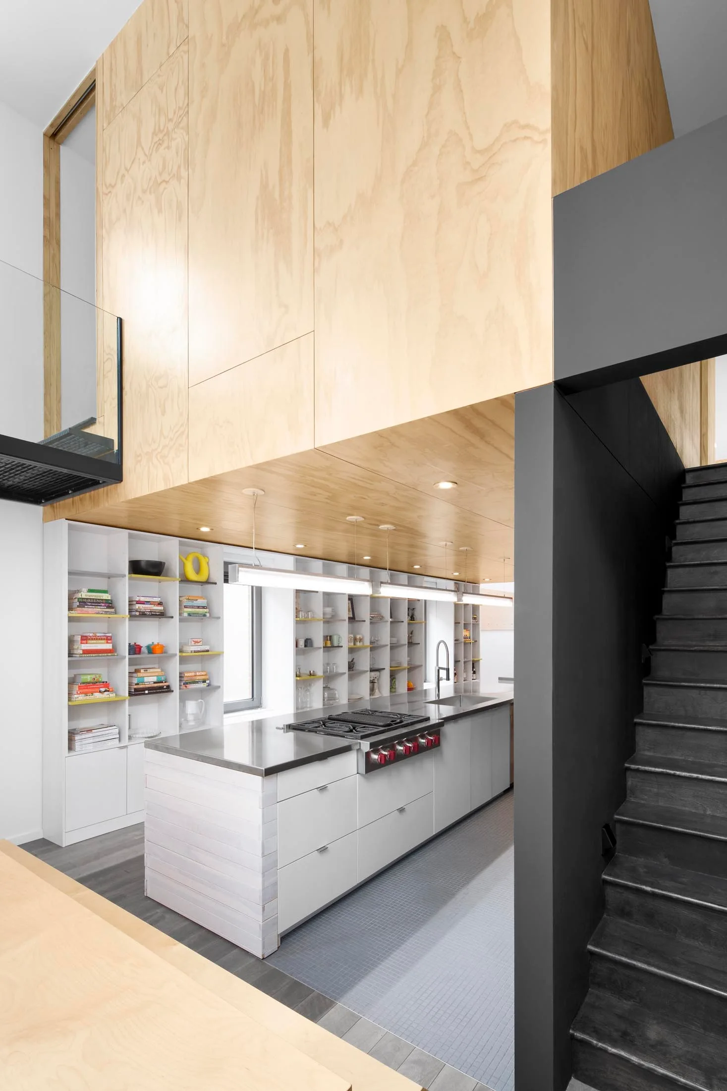 A minimalist kitchen features a stainless steel island below a plywood-clad ceiling, with white built-in bookshelves and a dark staircase.