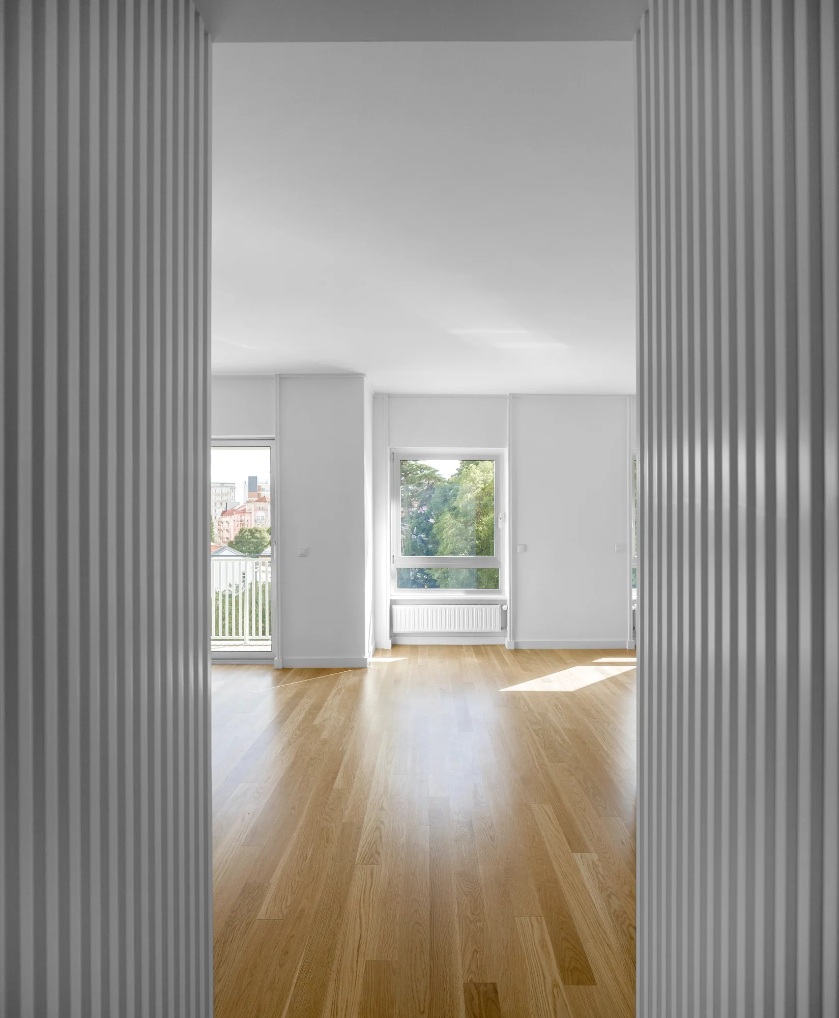 A view through white fluted doorway panels into a bright, empty room with light wood flooring and a window looking out at trees.