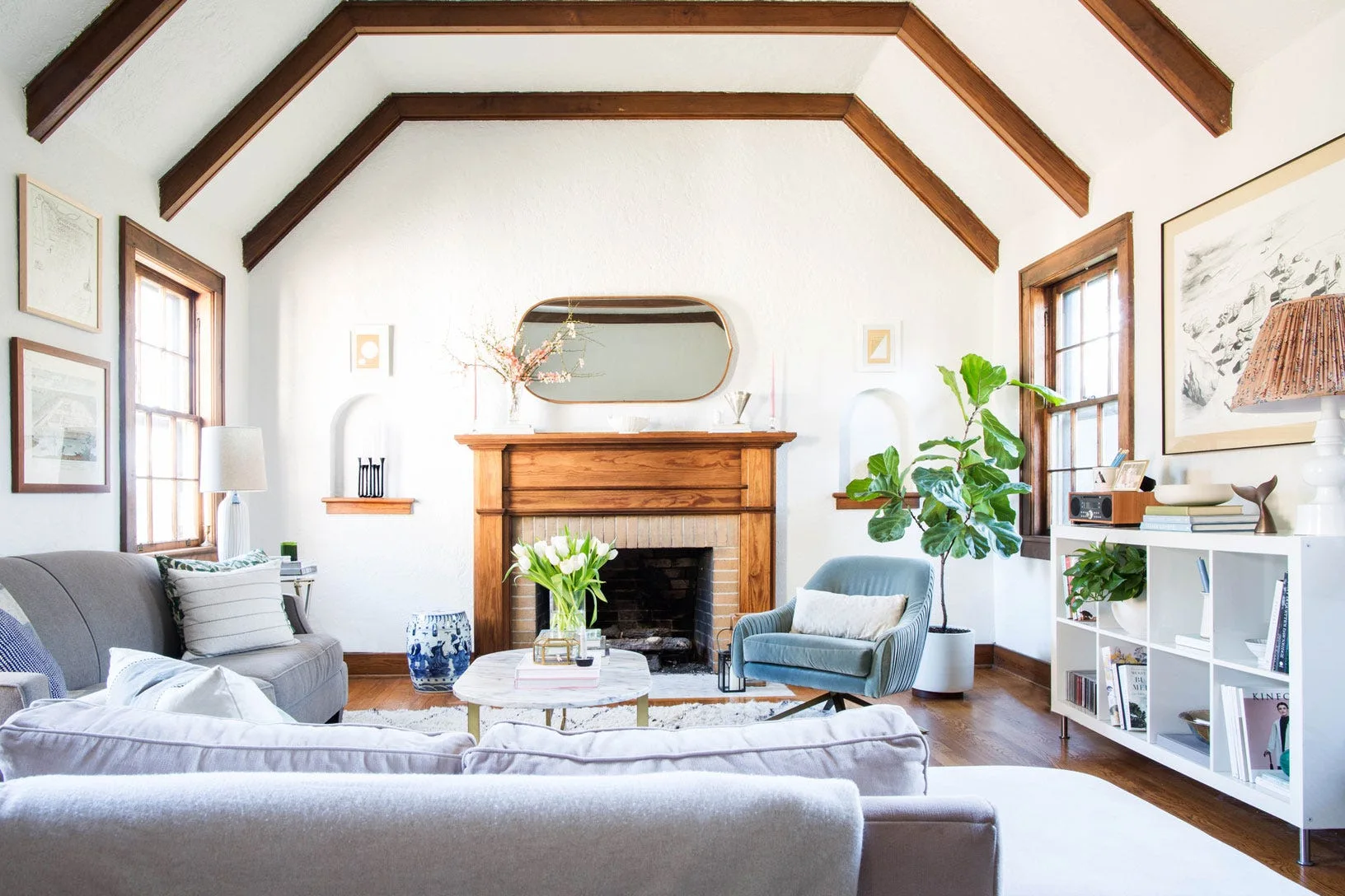 A bright living room with a vaulted wood-beamed ceiling, a central fireplace flanked by arched wall niches, a grey sofa, and a blue armchair.