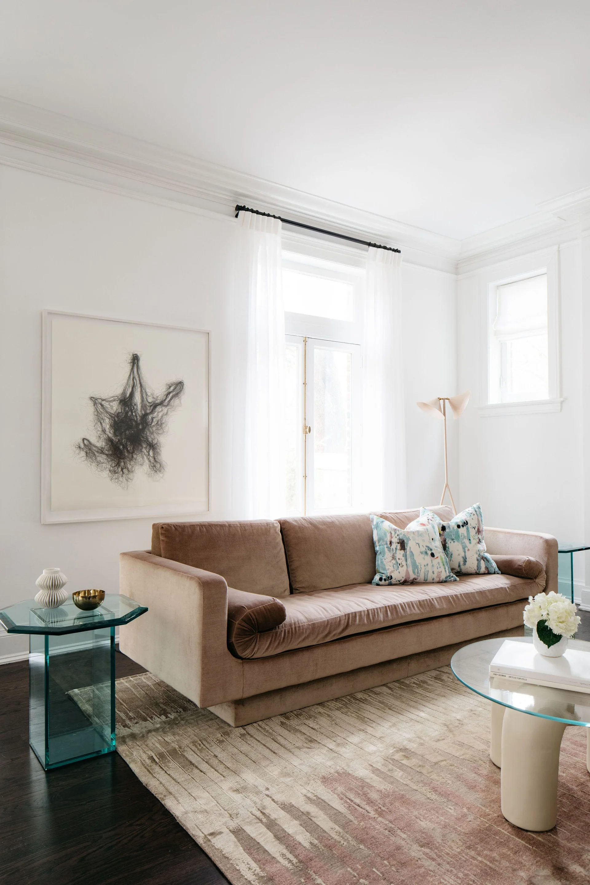 A beige velvet sofa sits on a patterned rug in a bright white living room with glass tables, sheer curtains, and abstract art.