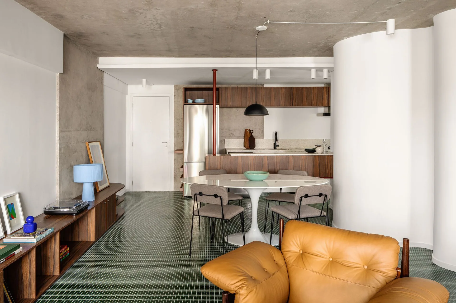 An open-concept room featuring a green tile floor, a round white dining table, a curved white wall, and kitchen cabinets below a concrete ceiling.
