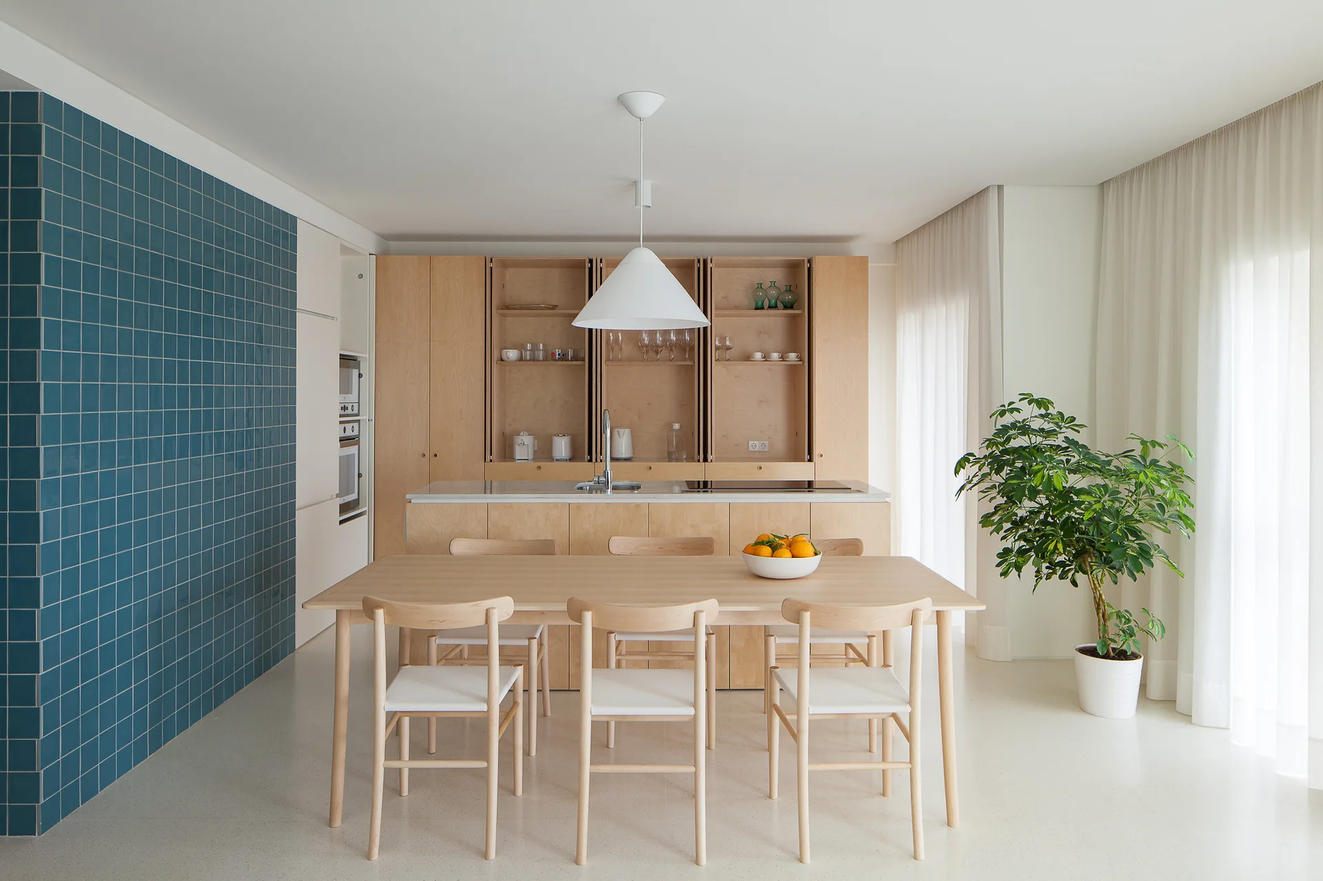 A minimalist kitchen and dining area features a blue tiled accent wall, light wood cabinetry, a dining table with chairs, and a white cone pendant light.