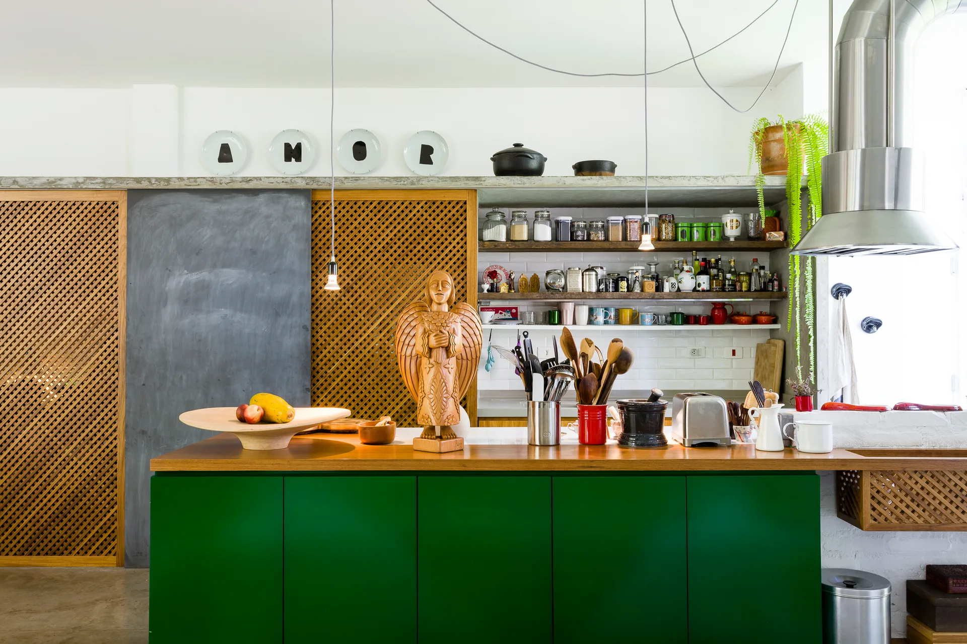 An eclectic kitchen features a forest green island with a wood countertop, open shelving stocked with jars, and a sliding wood lattice door.