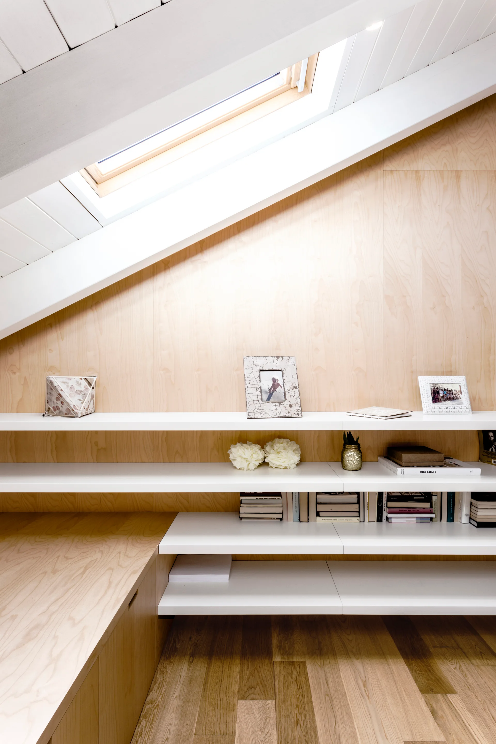 White built-in shelves are mounted against a light wood paneled wall in an attic room under a sloped ceiling with a skylight.