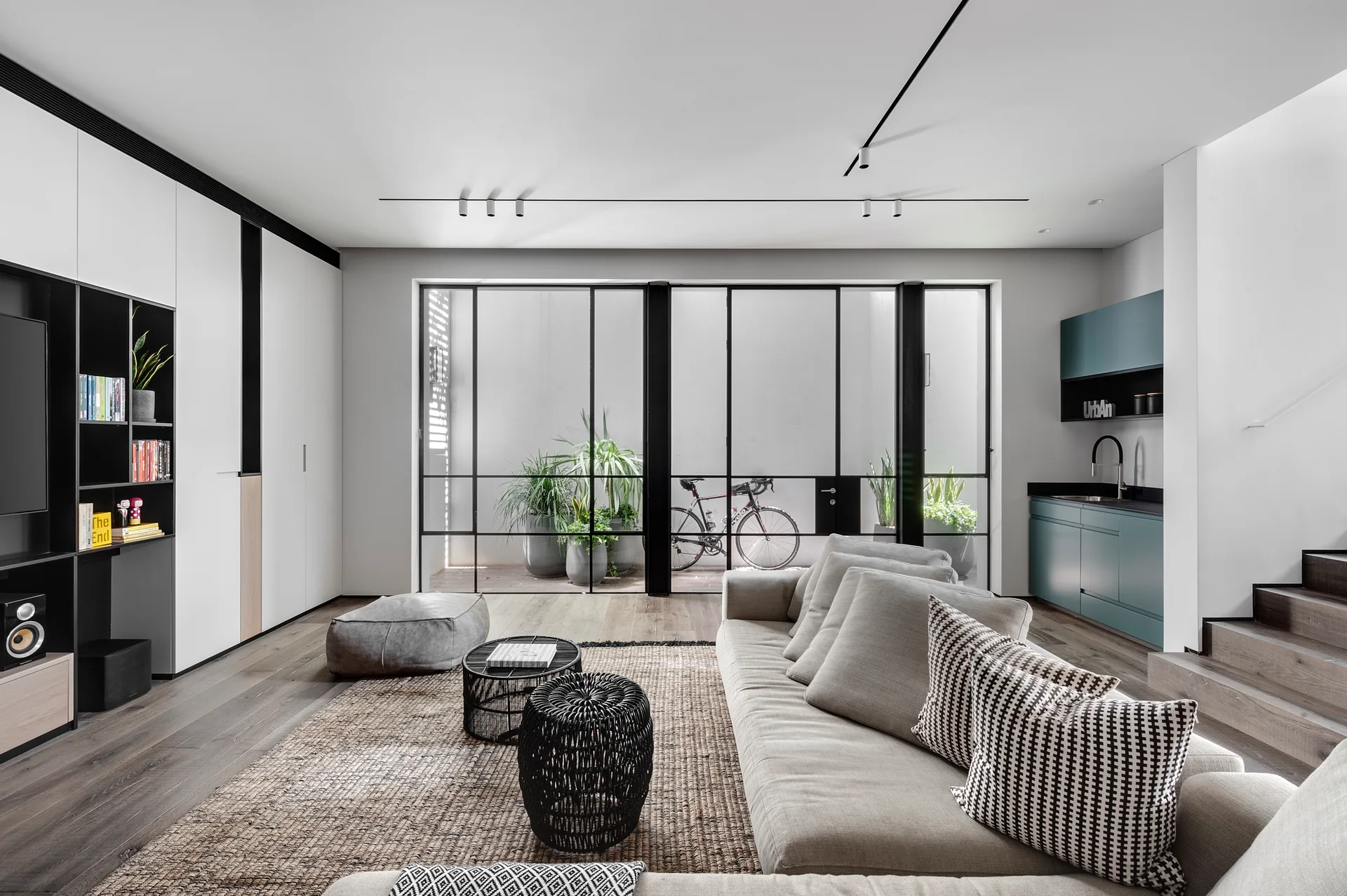A modern living room features a gray sofa on a woven rug, black track lighting on the ceiling, and black-framed glass doors leading to a terrace.