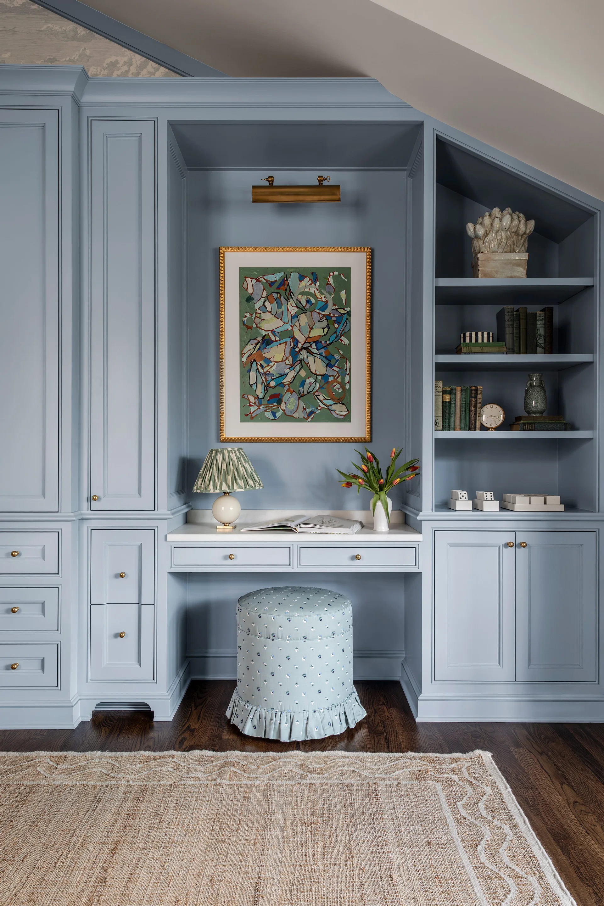 A light blue built-in desk unit with integrated cabinets, open shelving, a framed painting, and a matching skirted floral ottoman on a dark wood floor.
