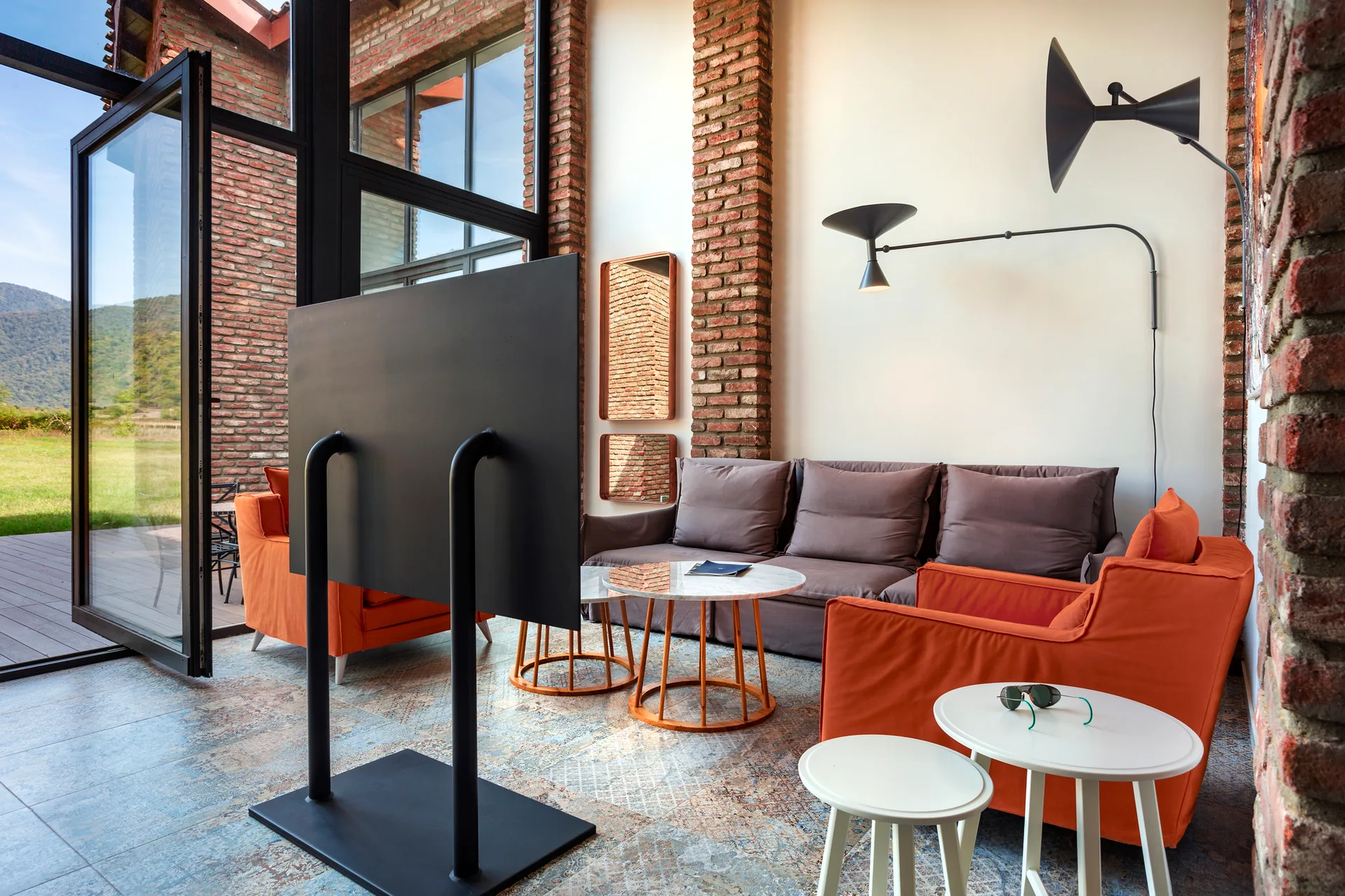A modern living room features a brown sofa, orange armchairs, exposed brick columns, and a TV on a black metal floor stand near large glass doors.