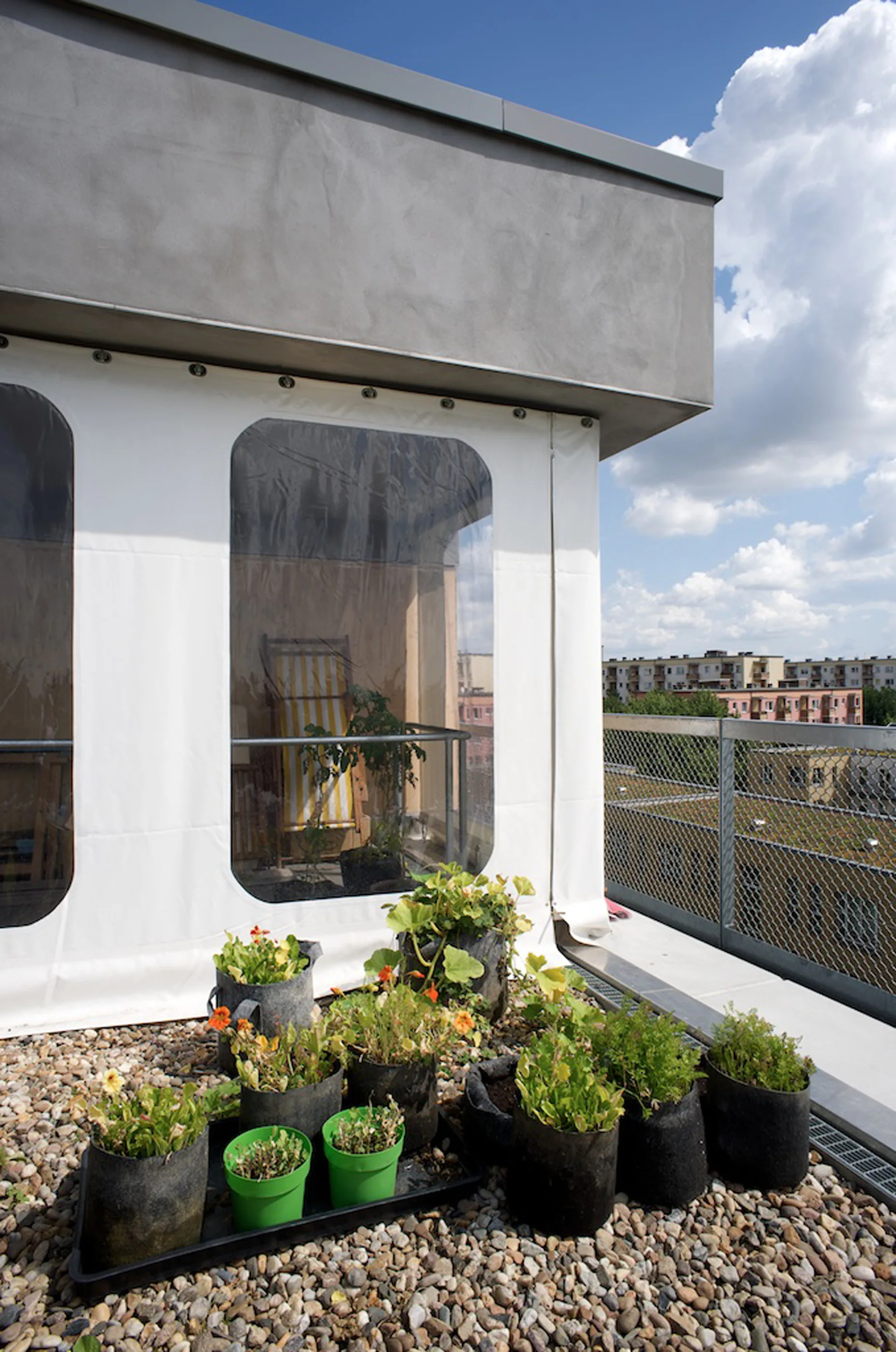 An elevated outdoor balcony featuring a pebble gravel floor, fabric container planters, a metal railing, and a clear vinyl enclosure.