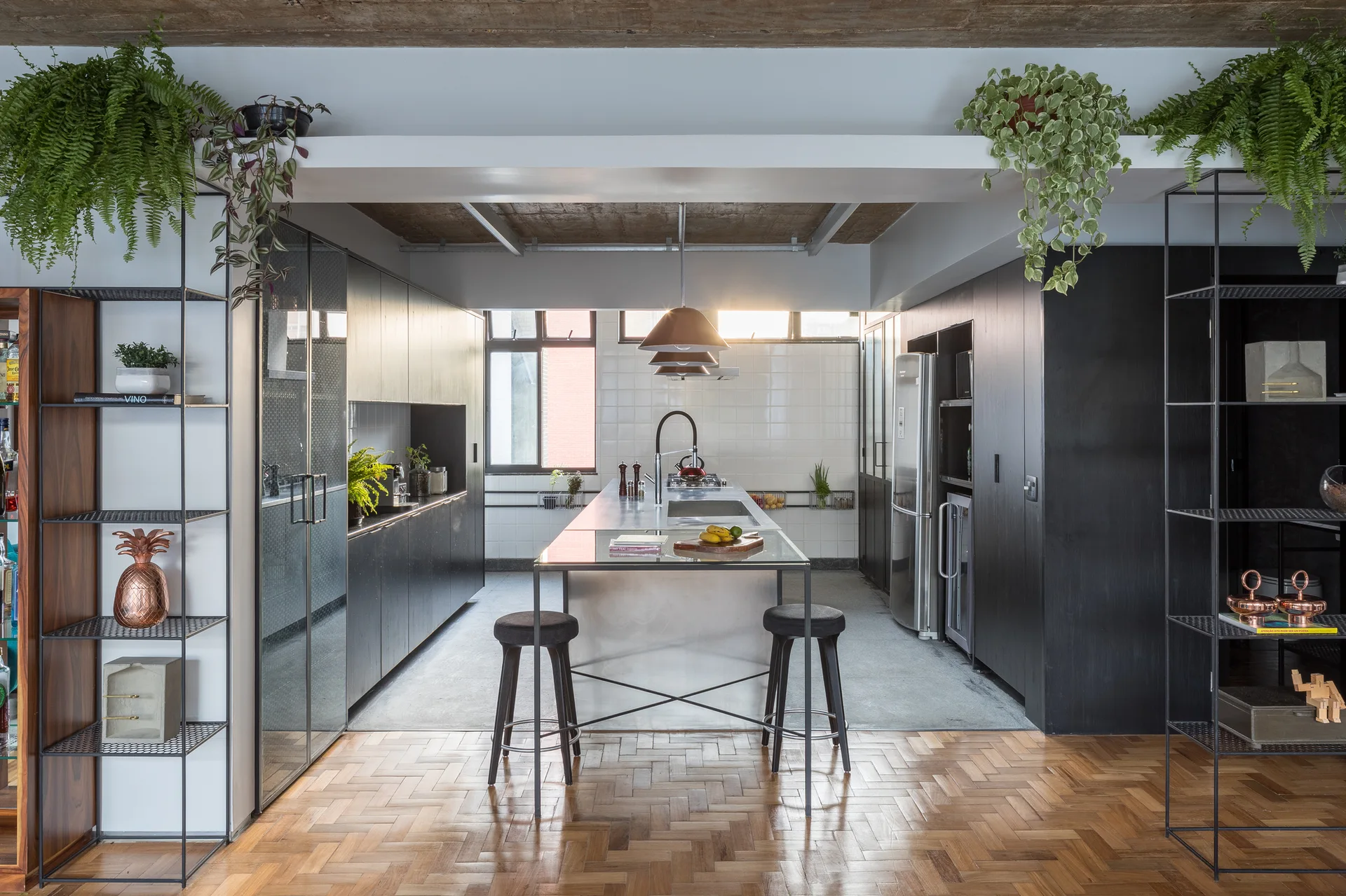 A modern kitchen featuring a stainless steel central island with stools, matte black cabinets, herringbone wood floors, and trailing green plants.
