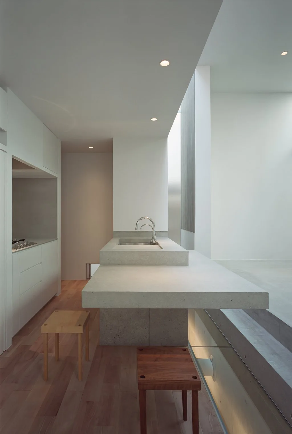 A minimalist kitchen featuring a cantilevered concrete island with an integrated sink, white flat-front cabinetry, and wooden stools.