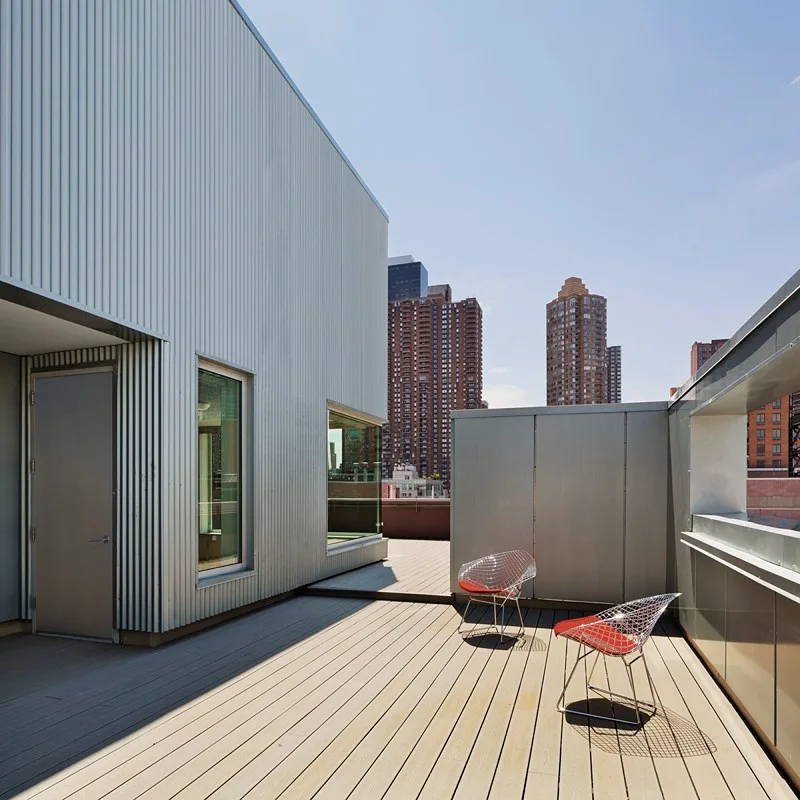 A modern rooftop terrace features corrugated silver metal siding, wood decking, and two metal wire chairs with bright red cushions against a city skyline.