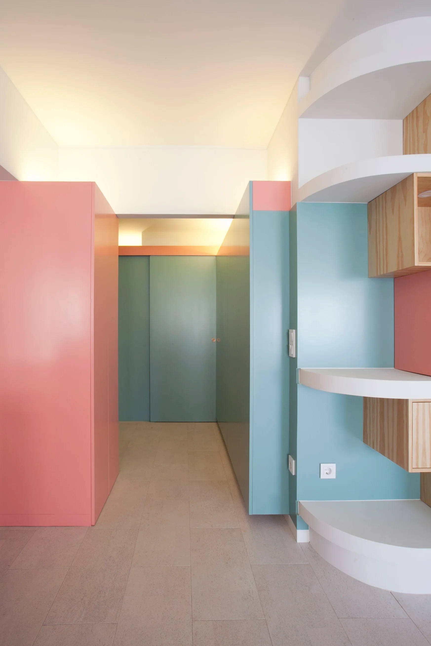 A modern hallway features matte pink and blue built-in storage cabinets, white curved open shelving, and warm indirect cove lighting.