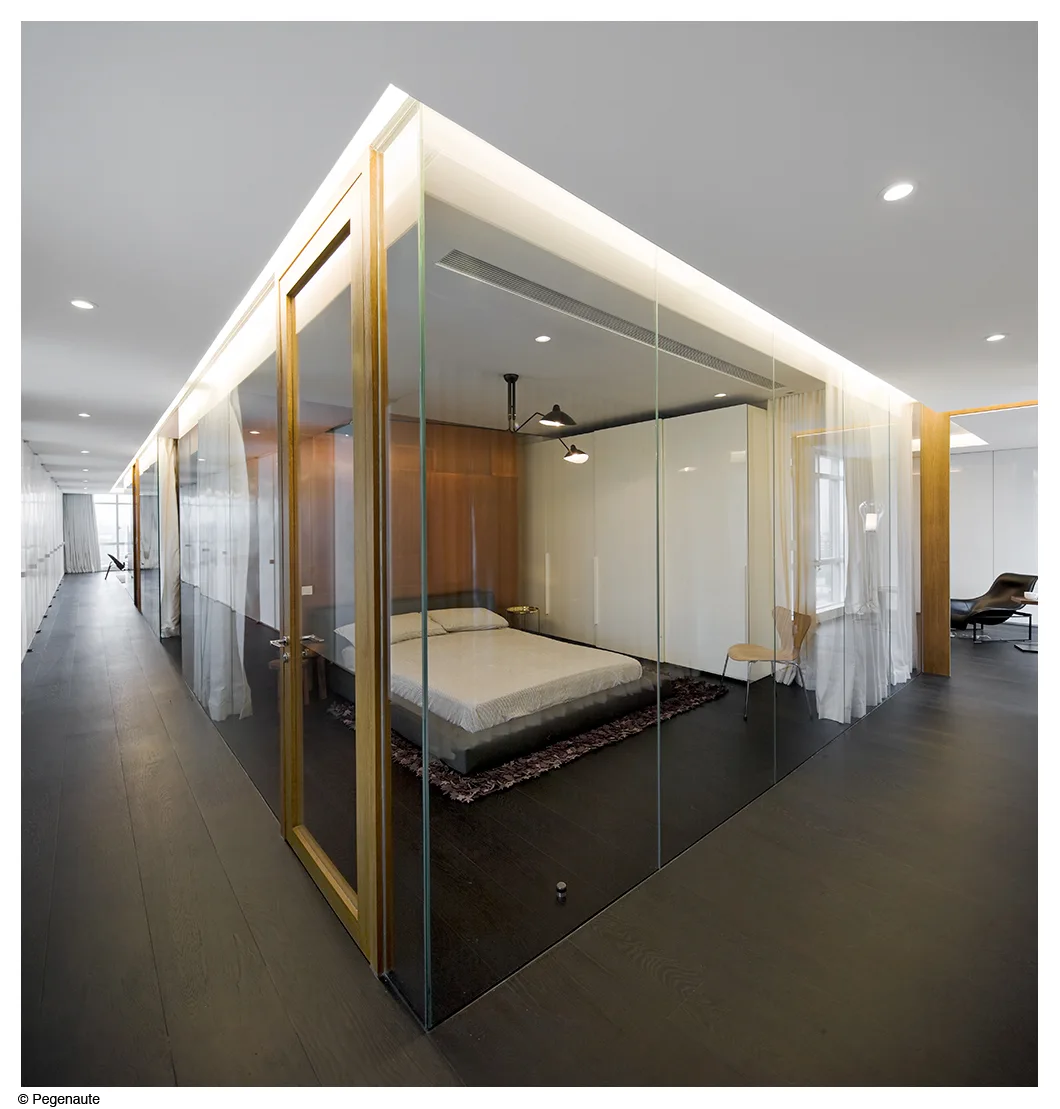A minimalist bedroom enclosed in frameless glass walls sits on a dark wood floor with built-in wardrobes and sheer curtains.