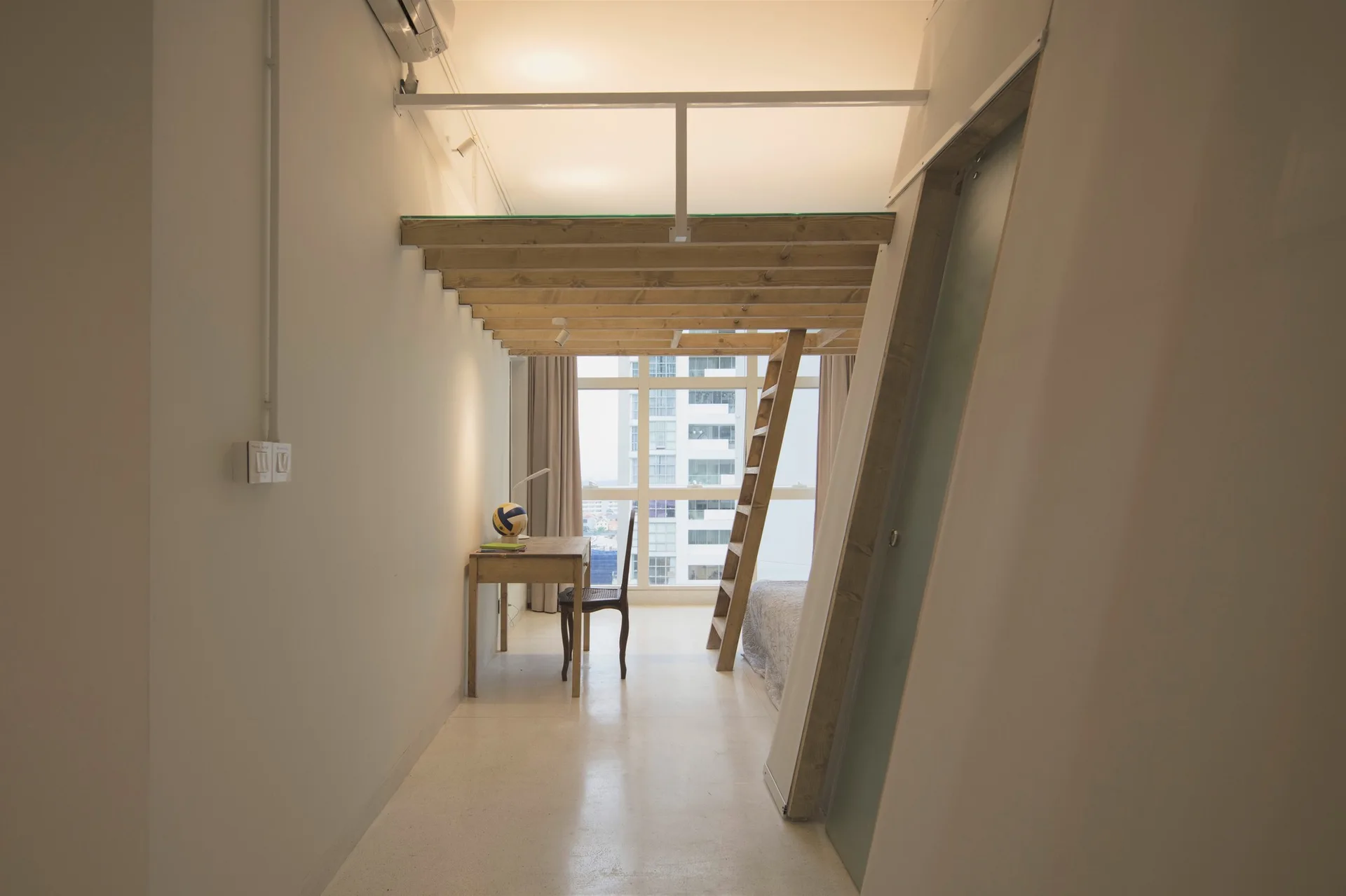 A minimalist white room features a light wood loft bed with a ladder, a simple writing desk, and a large window facing city buildings.