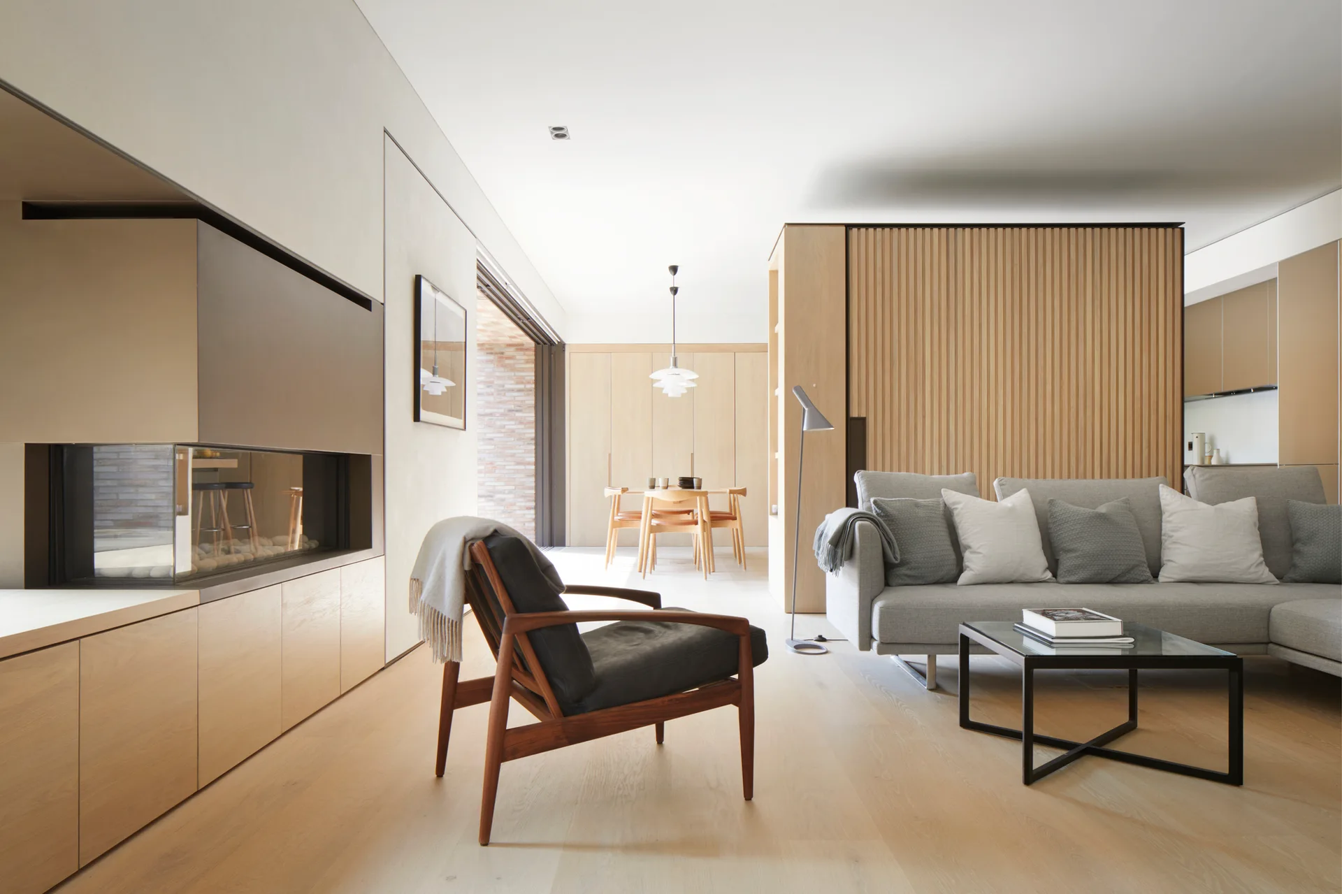 A minimalist open-plan living room with a grey sofa, vertical wood slat wall, leather armchair, and built-in modern fireplace.