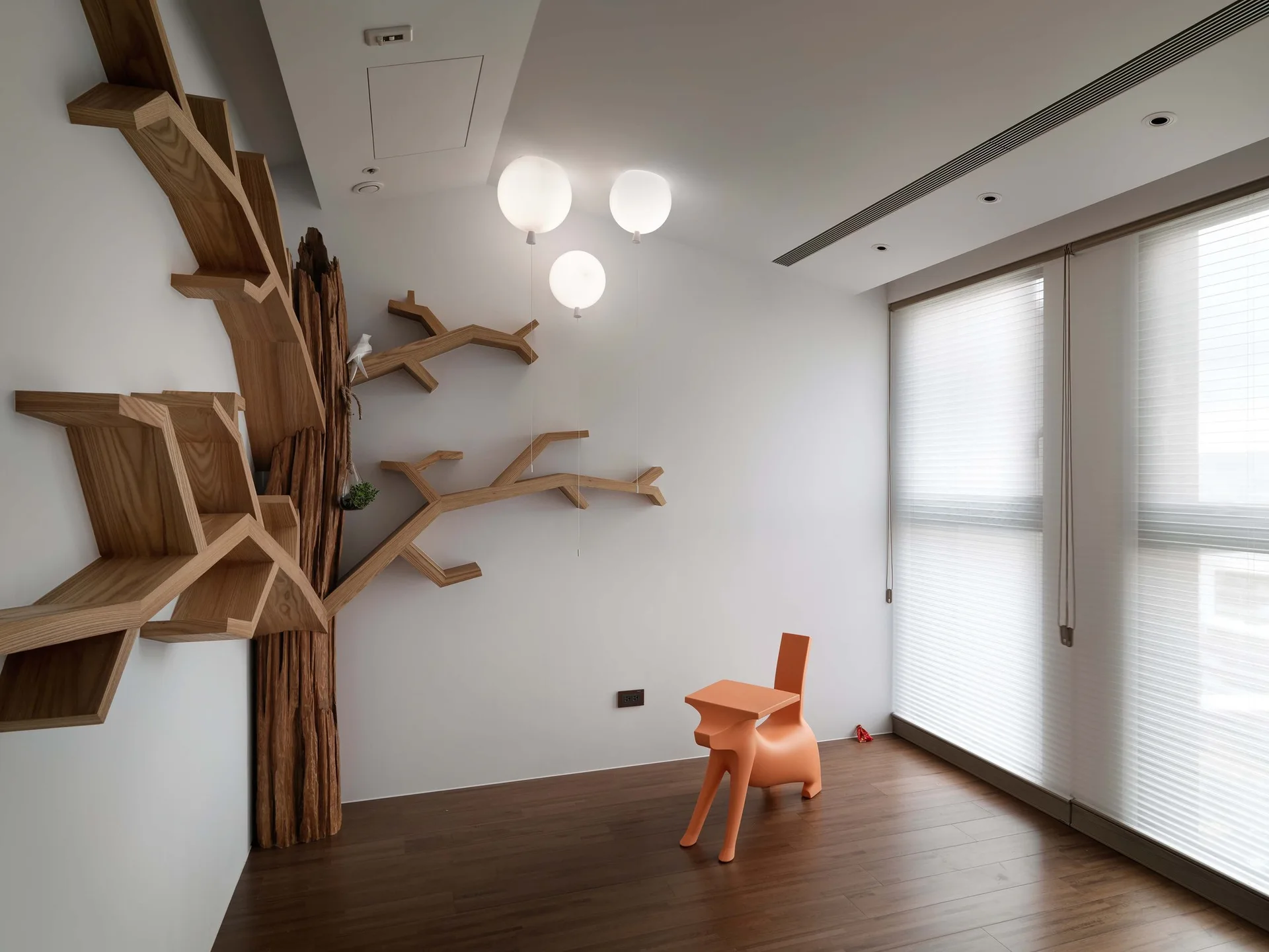 A kids playroom features a sculptural wooden tree bookshelf on a white wall and a bright orange animal-shaped chair.
