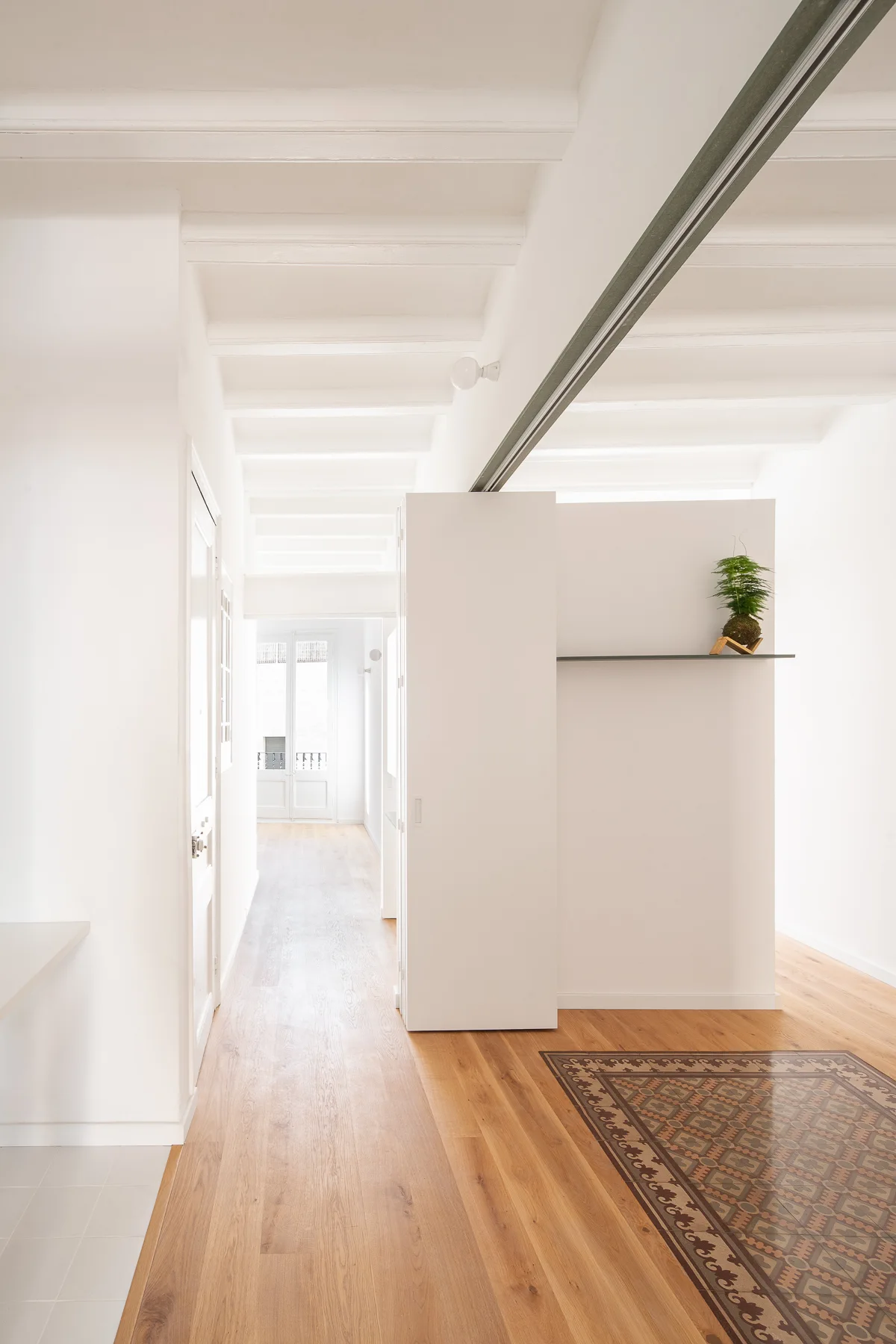 A bright hallway featuring light wood flooring with a patterned tile inlay, white walls with exposed ceiling joists, and a central partition.