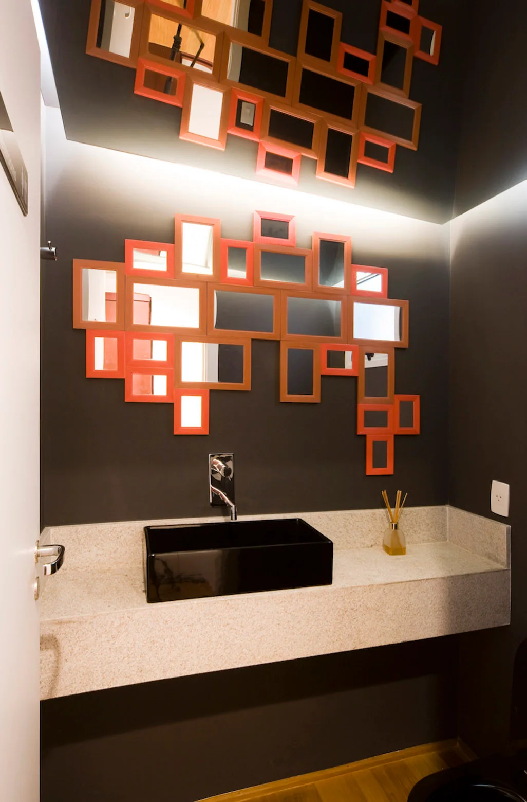 A black rectangular vessel sink sits on a beige stone floating vanity against dark walls decorated with a cluster of orange-framed mirrors.