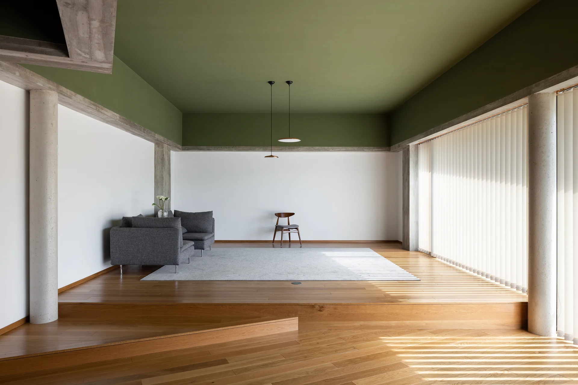 A minimalist living room features a grey sofa on a raised wooden platform, concrete pillars, and an olive green ceiling.