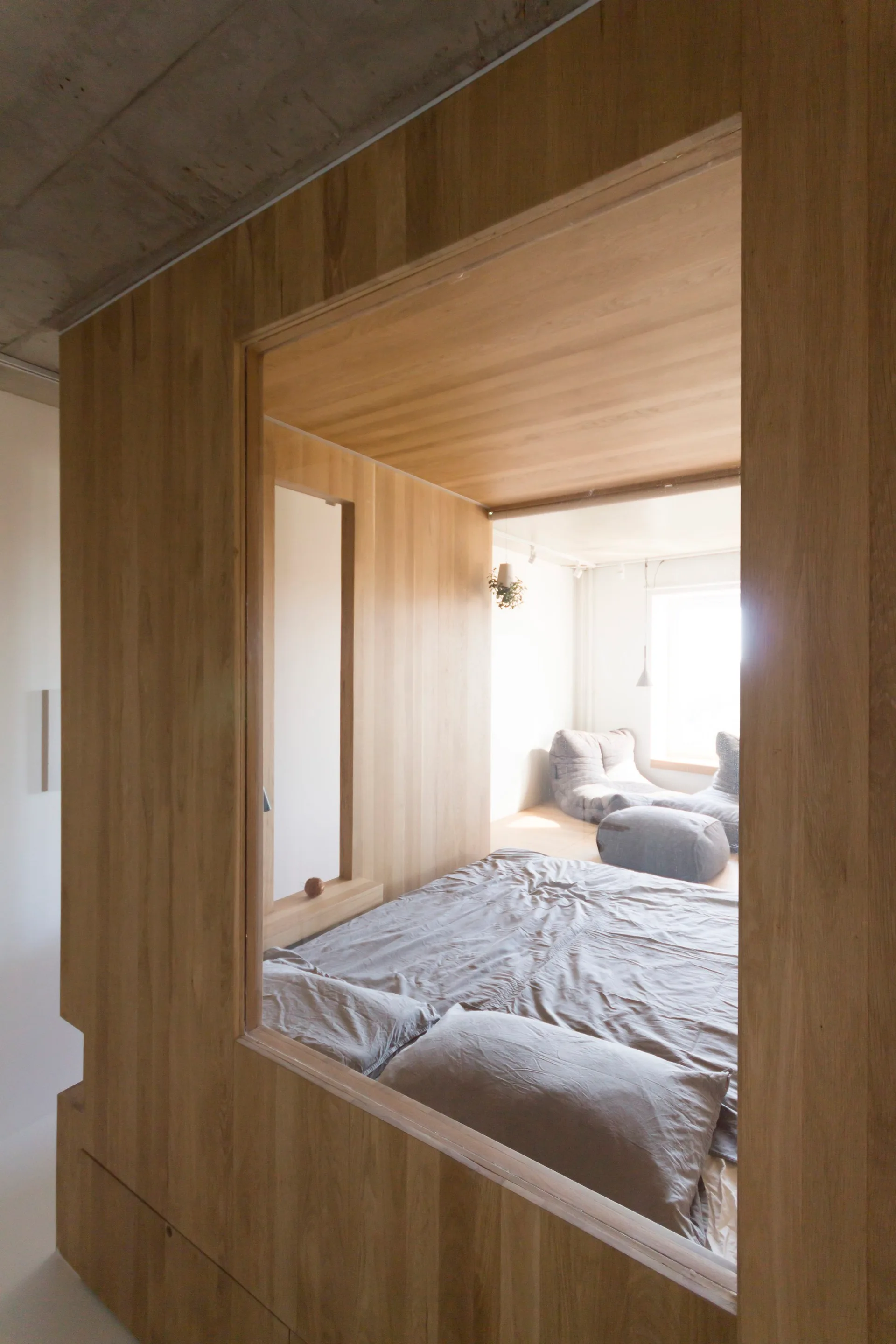 A low-profile bed with grey linens is enclosed inside a custom wooden wall partition featuring a large glass window.
