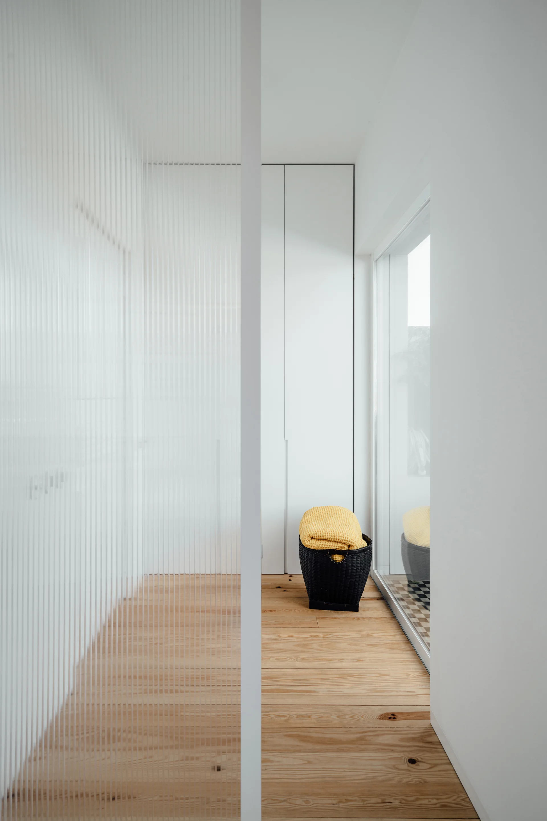 A minimalist white hallway features a fluted glass room divider, light wood flooring, tall white cabinets, and a black basket with a yellow blanket.