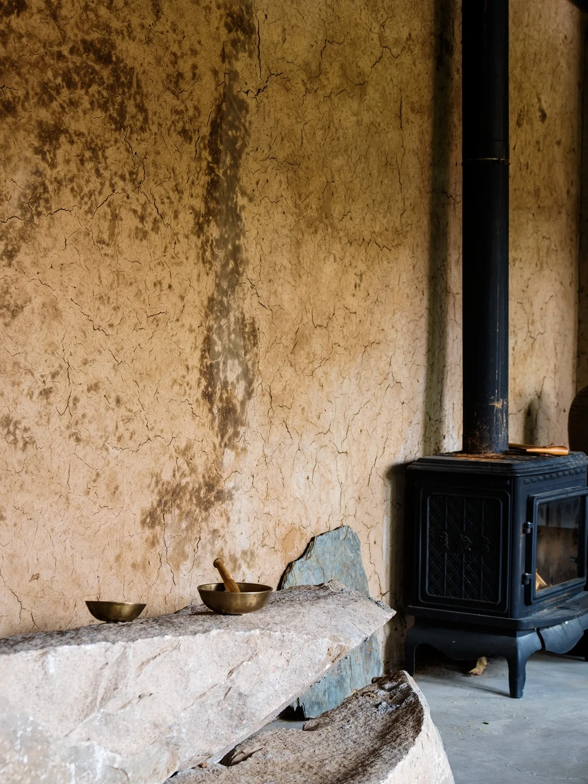 A rustic room features a cracked clay plaster wall, a black cast iron wood stove, and a rough-cut natural stone bench holding two brass bowls.