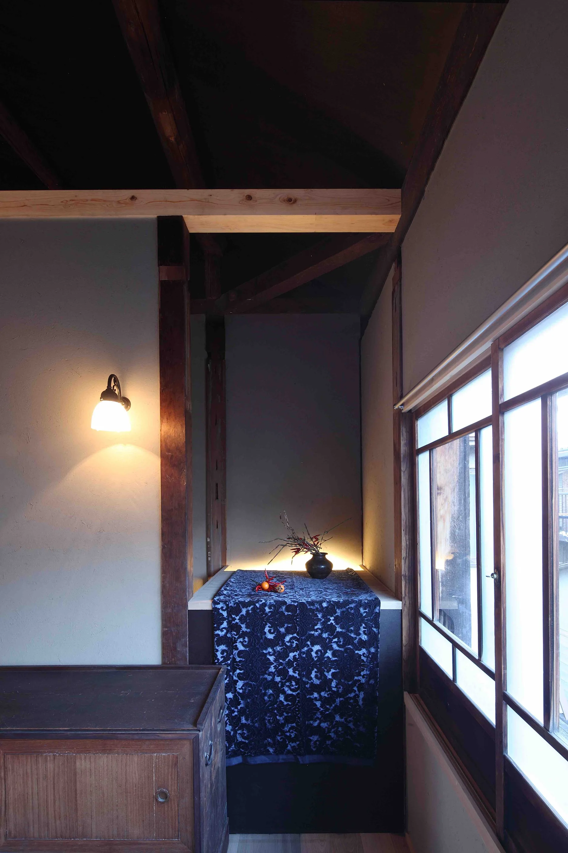 A dimly lit interior with traditional Japanese wood beams, a wall sconce, and a recessed alcove decorated with a patterned blue runner.