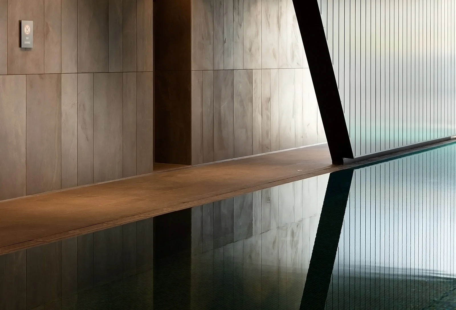 A minimalist indoor swimming pool with vertical wood-paneled walls, a flat stone pool deck, and clear reflections in the calm water.