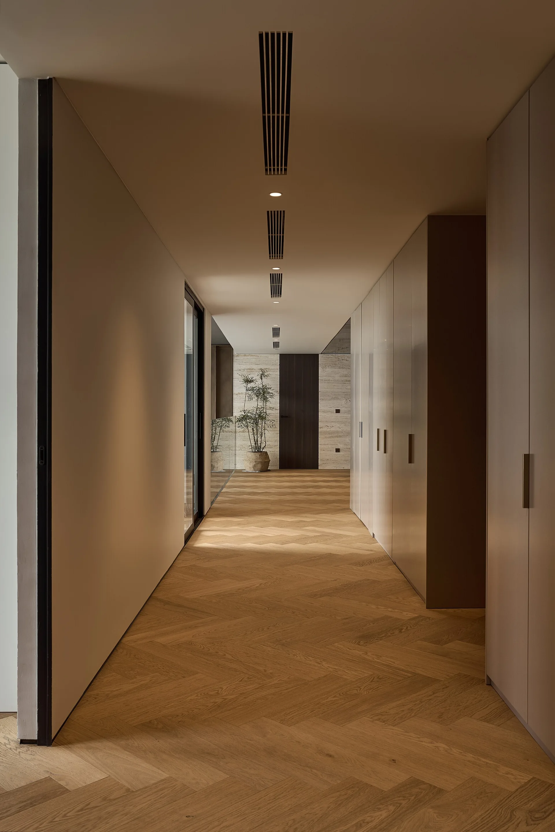 A long minimalist hallway features herringbone-patterned oak flooring, floor-to-ceiling taupe storage cabinets, and linear recessed ceiling vents.