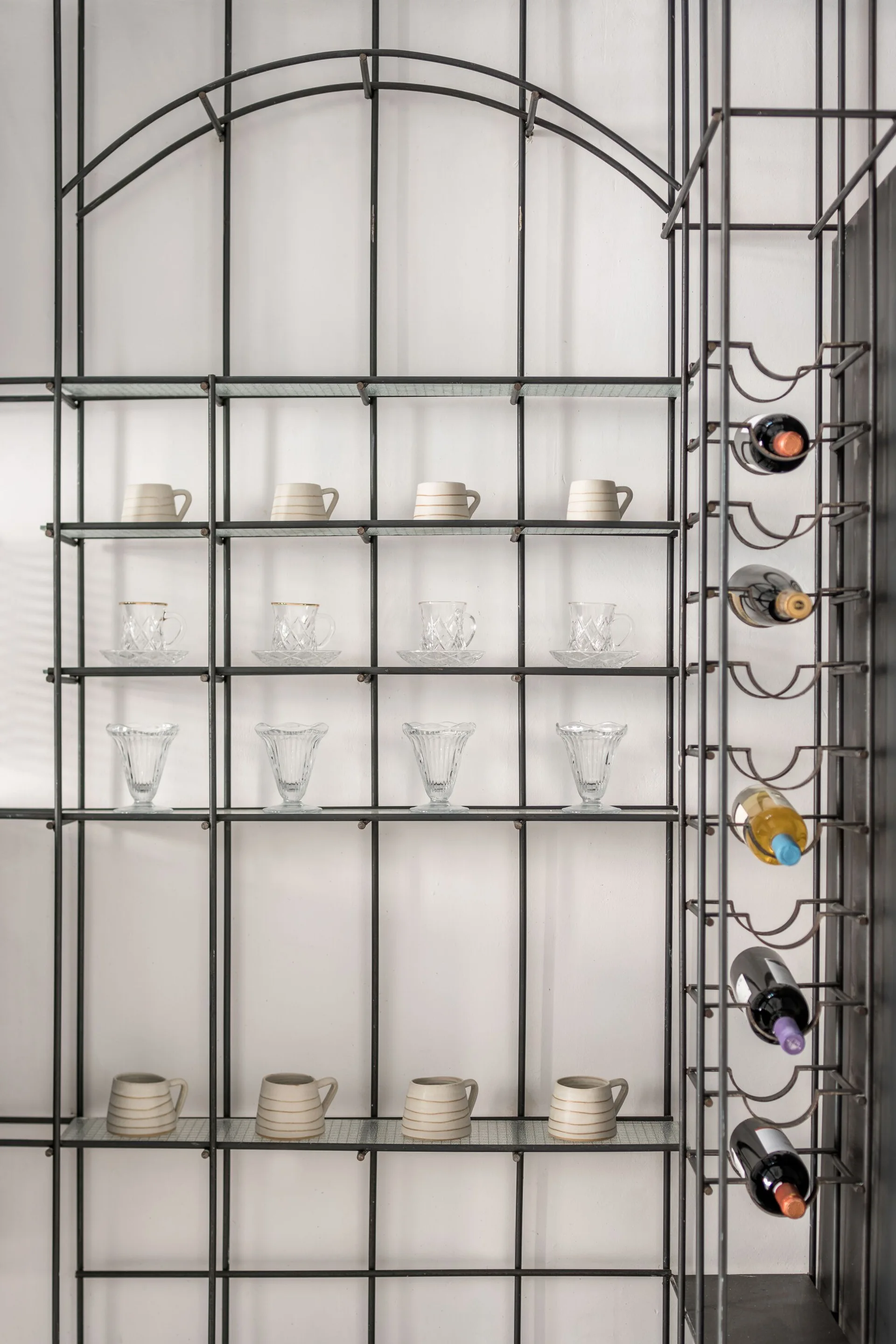 A black metal grid shelving unit with an arched top holds rows of ceramic cups and clear glassware next to an integrated wine rack on a white wall.