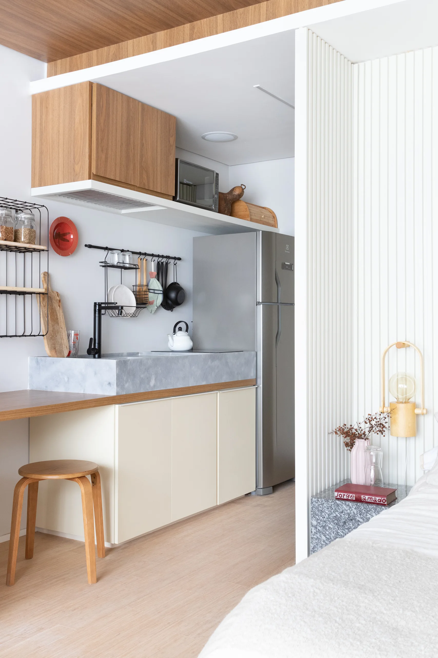 A compact kitchenette with light beige cabinets and a grey stone sink sits next to a sleeping area with vertical ribbed white wall paneling.