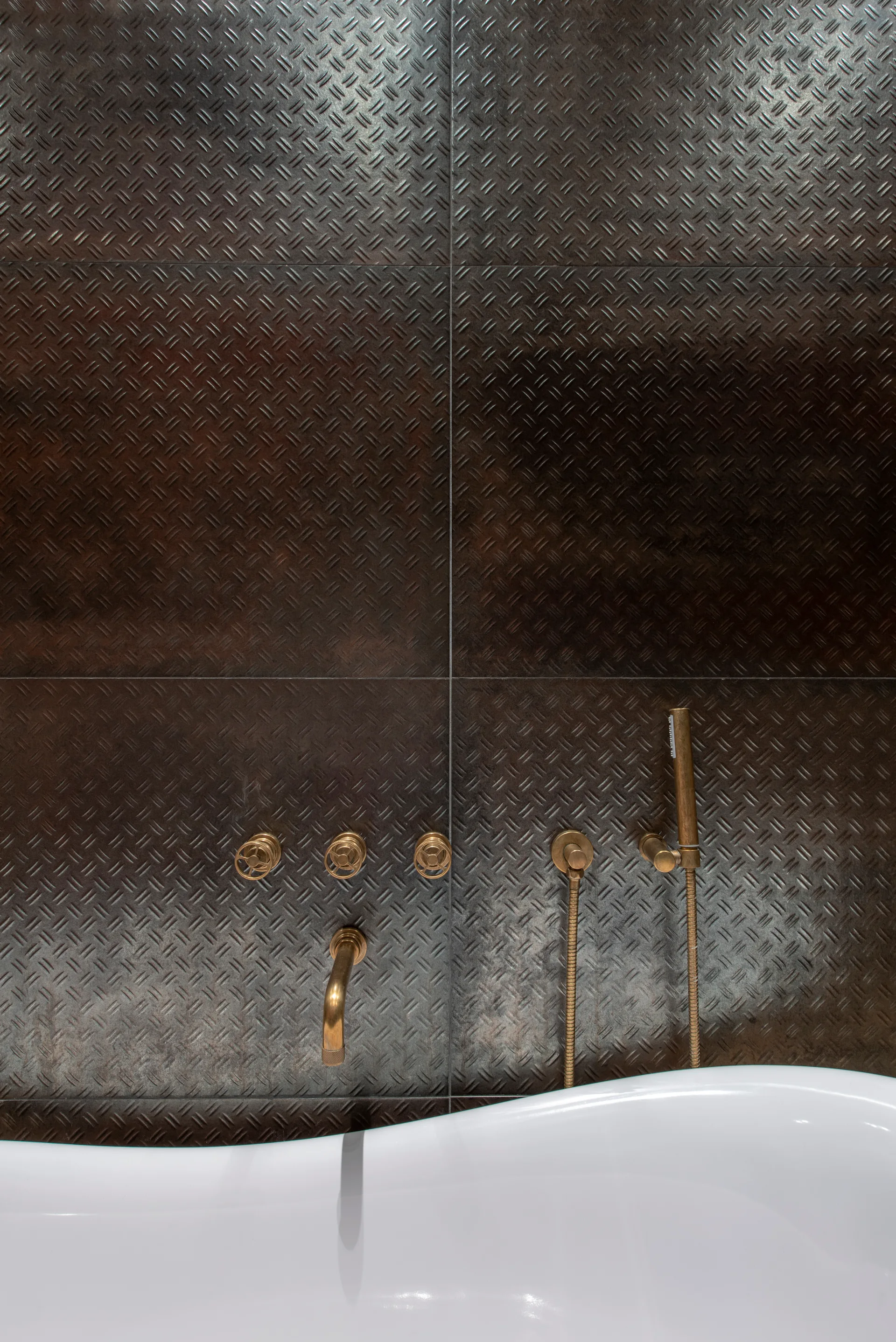 A white ceramic bathtub sits against a dark gray metal diamond plate wall featuring wall-mounted brushed brass plumbing fixtures and a hand shower.