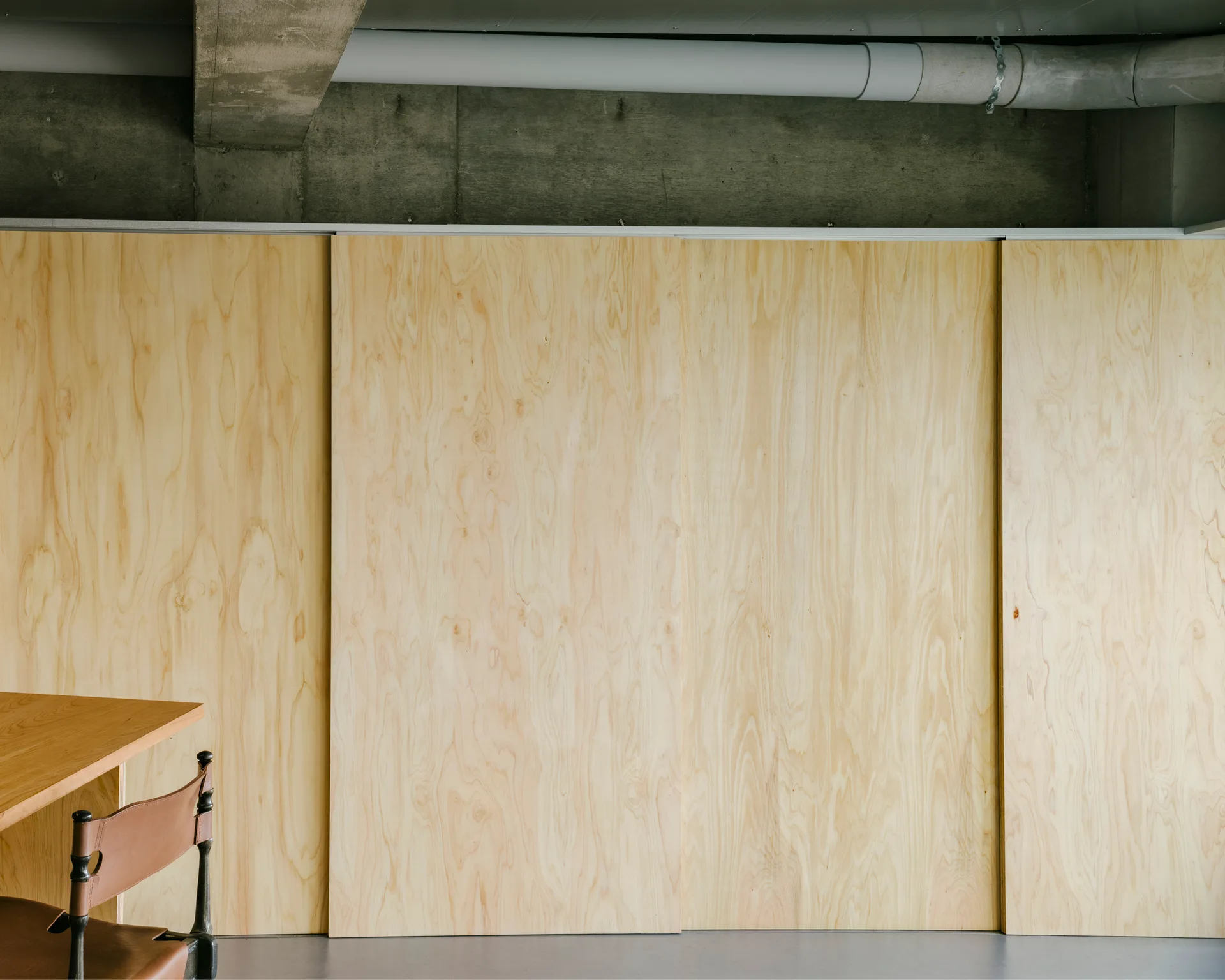Large plywood sliding panels cover a wall below a raw concrete ceiling with an exposed utility pipe and concrete beam.