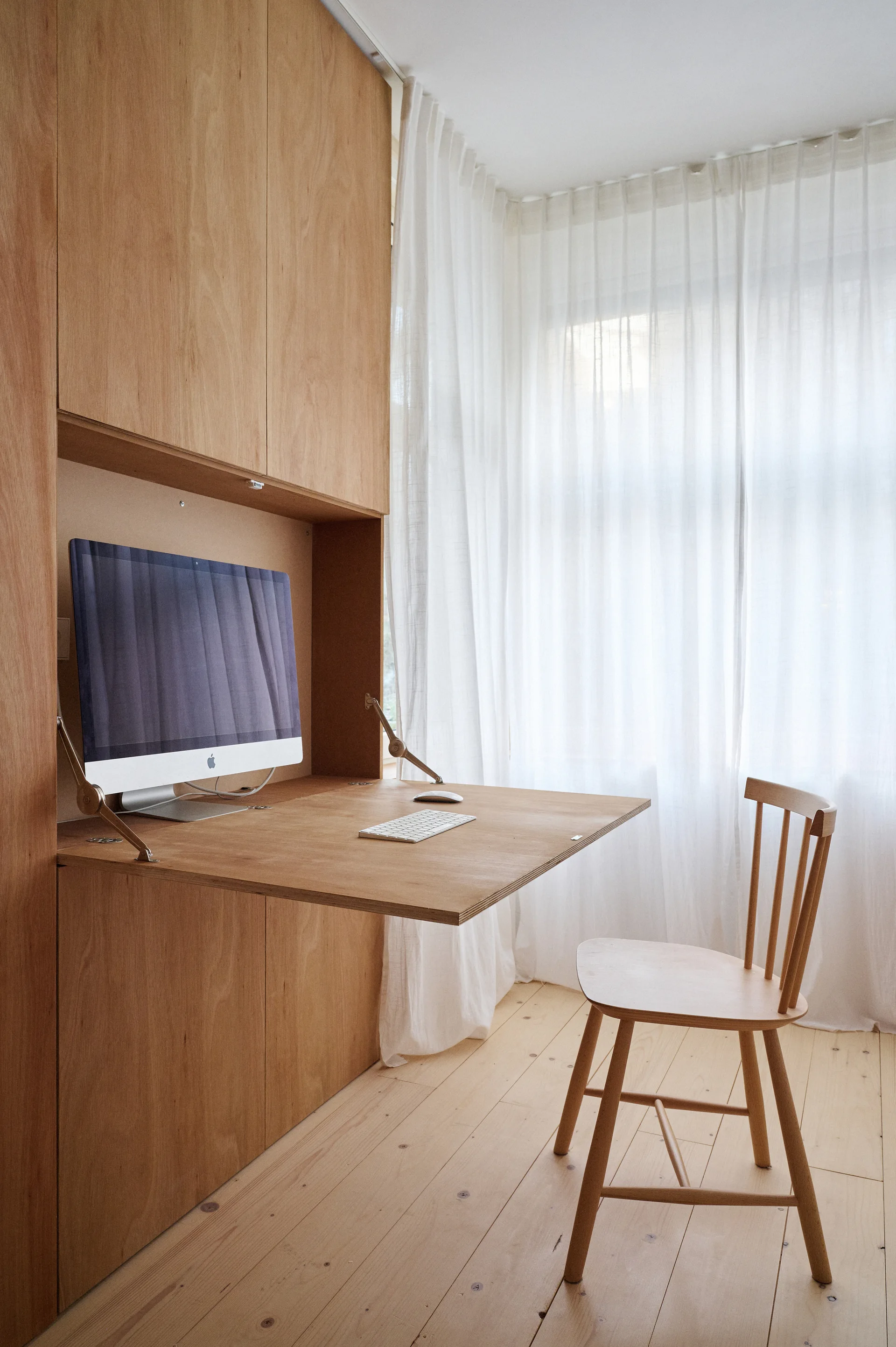 A minimalist home office features a built-in light wood cabinet with a fold-down desk, a wooden spindle chair, and sheer white curtains.