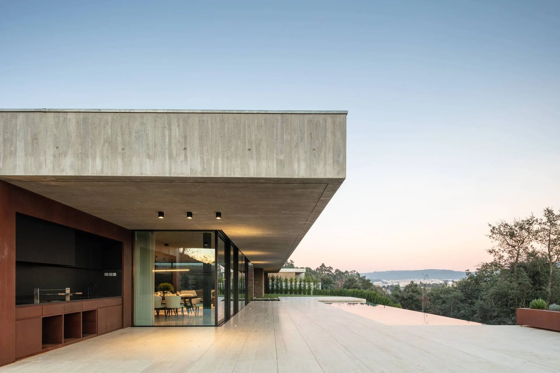 A concrete terrace featuring a rust-colored outdoor kitchen, large stone floor tiles, and an infinity pool under a cantilevered concrete ceiling.