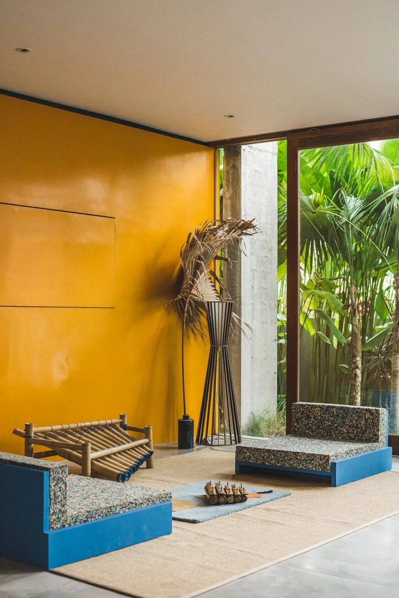 A modern living room features a mustard yellow accent wall, low blue chairs with speckled foam cushions, and a large glass door framed by tropical palms.