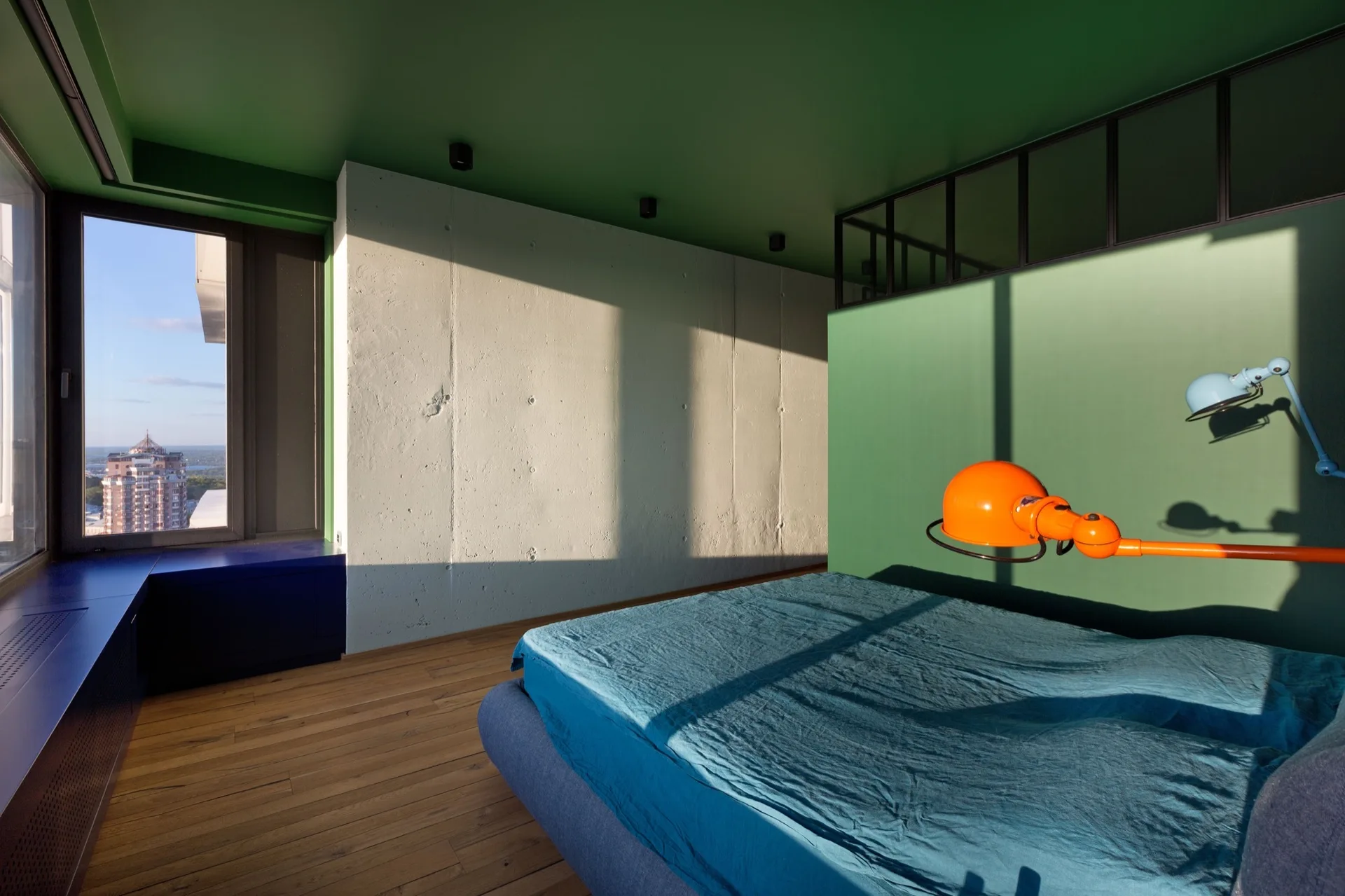 Modern bedroom with a green painted ceiling and wall, a raw concrete accent wall, a blue bed, and a colorful orange swing-arm lamp.