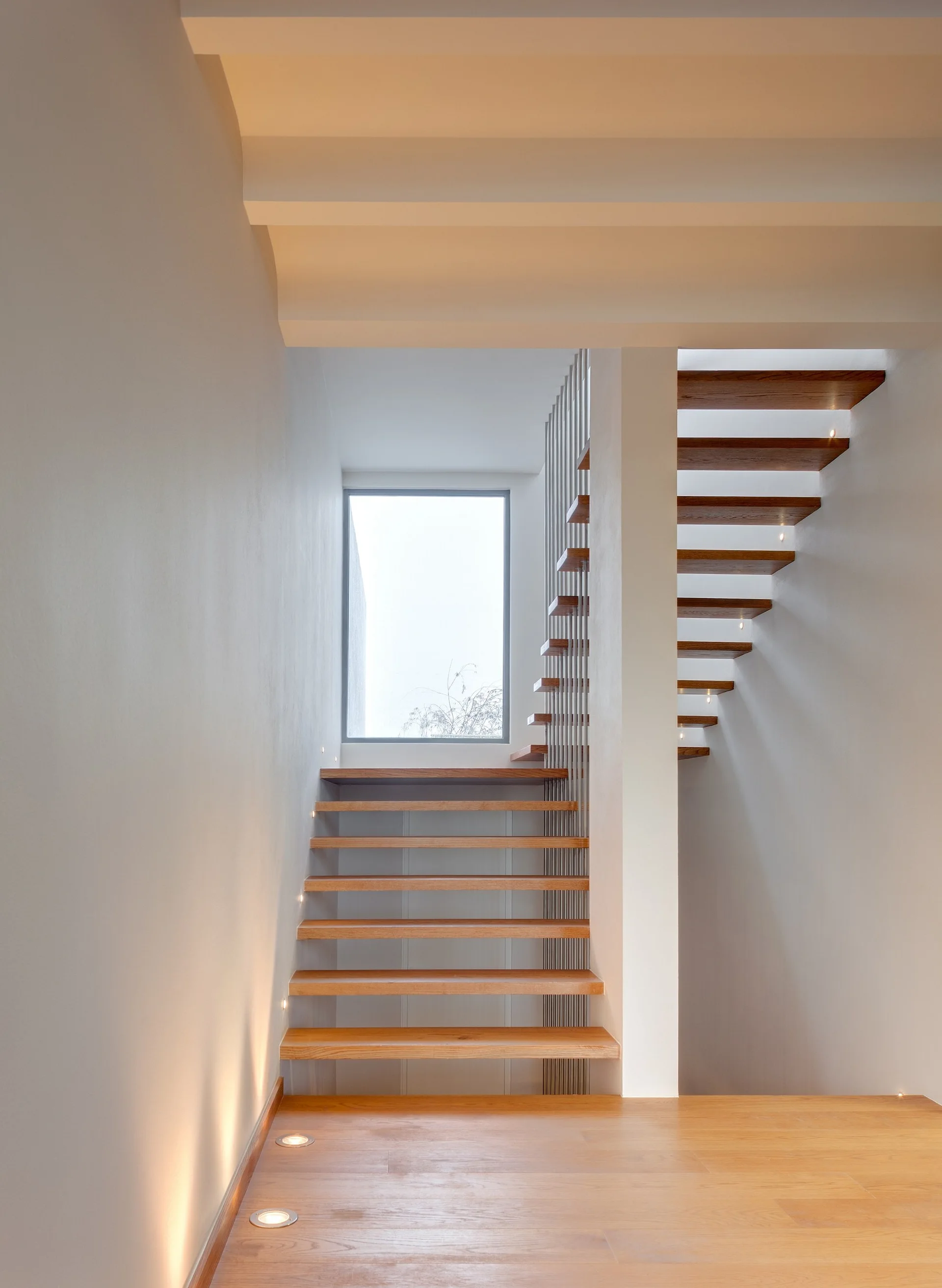 A modern floating wooden staircase with vertical metal rod balusters, recessed wall lighting, and a square window on the white wall landing.