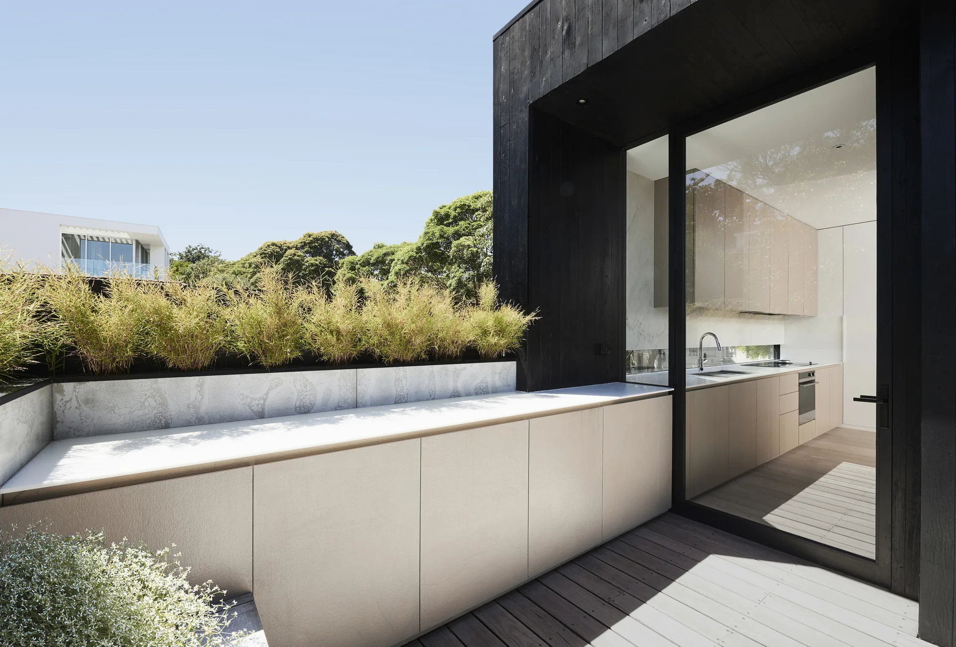An outdoor deck features light beige cabinetry with an integrated planter next to a black charred-wood wall and a sliding glass door.