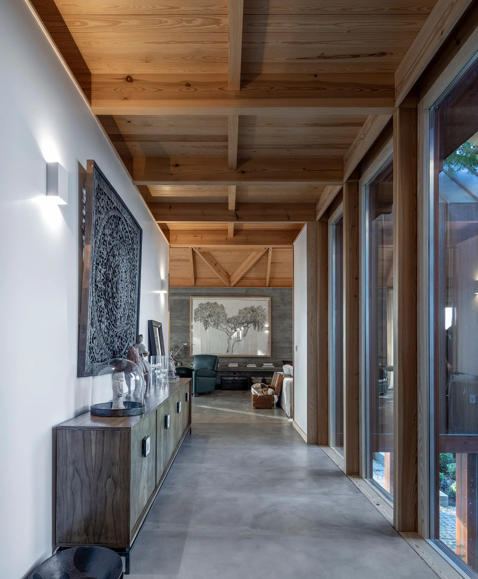 A modern residential hallway featuring a polished concrete floor, a wood plank ceiling with beams, a wood sideboard, and floor-to-ceiling glass windows.