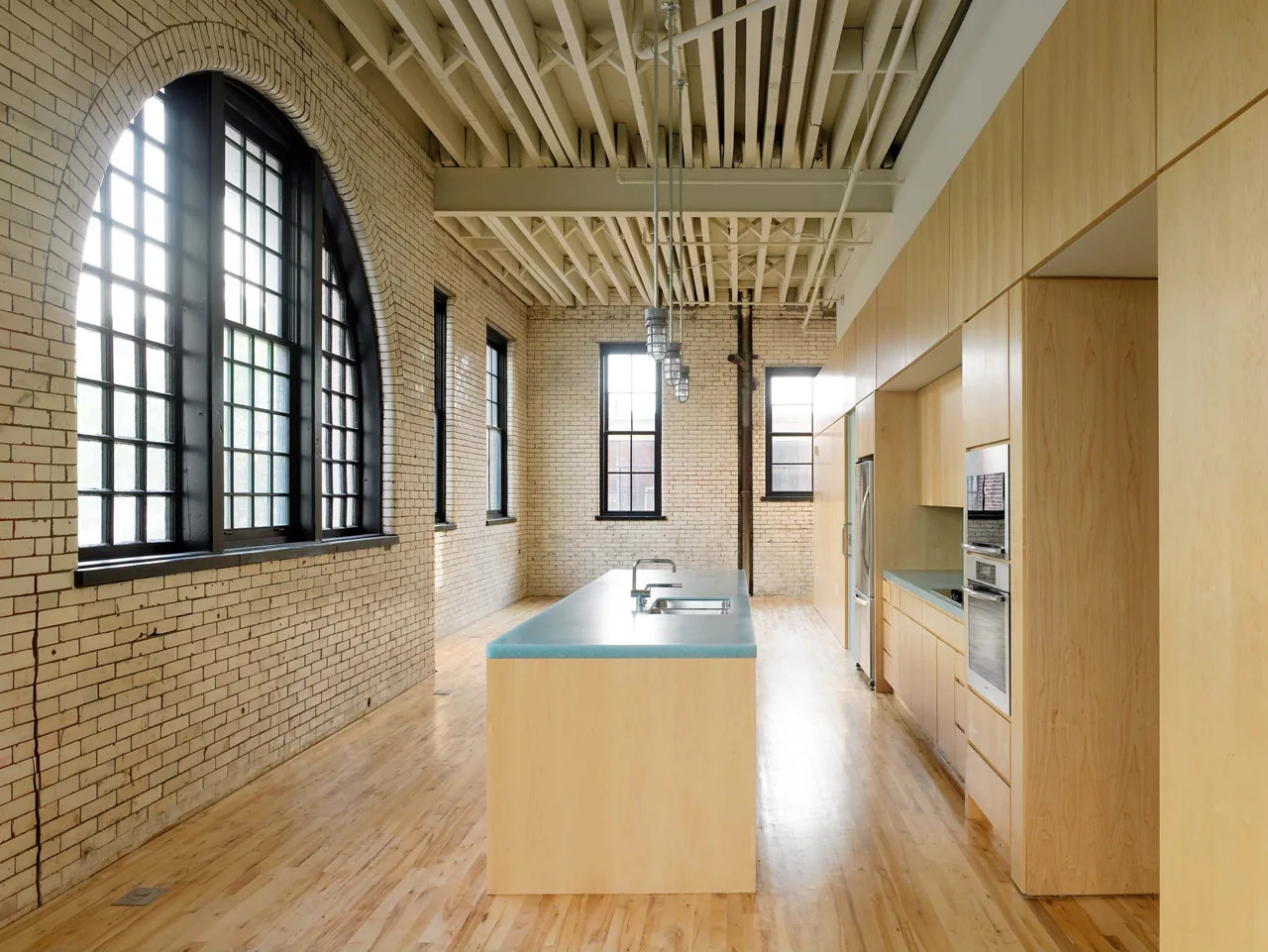 A spacious loft kitchen features light wood cabinets, an island with a blue countertop, off-white brick walls, and a large arched window.